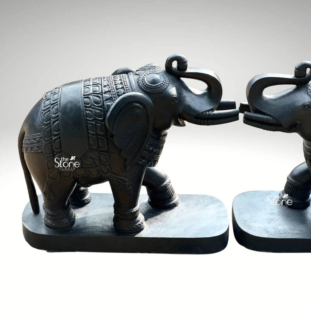 A close-up three-quarter view photograph captures a magnificent pair of 26-inch blackstone elephant statues meticulously hand-carved and arranged with their trunks touching in a gesture of affection and greeting. The elephants are crafted from dark rich blackstone giving them a smooth polished texture that highlights the intricate details of their form. Each elephant is adorned with finely etched patterns on their backs resembling ornate decorative blankets and has detailed carvings on their legs ears and trunks showcasing a high level of traditional Indian craftsmanship. The elephants are depicted with their trunks curled and interlocked a pose symbolizing good luck strength and prosperity common in Indian elephant iconography. The pair of statues is positioned symmetrically on a simple unadorned blackstone base, which grounds the monumental figures. The elephants' substantial 26-inch length makes them an imposing and inspiring presence suitable for a grand home interior entryway or living space consistent with the article's theme of a blackstone elephant statue pair. The dark stone material provides a powerful and elegant contrast against a clean neutral light-grey background allowing every nuance of the carving to be appreciated. Even directional studio lighting illuminates the pair emphasizing the deep textures and sharp lines of the carvings across the dark polished surface of the stone. This image is an exceptional representation of a traditional blackstone elephant statue pair showcasing the rich cultural artistry and spiritual significance of such a piece. The fine details from the delicate patterns on the decorative blankets to the lifelike form of the raised and touching trunks and flowing tails demonstrate the artisan's skill in working with this durable and classic medium. The statues' posture with the elephants facing each other with touching trunks conveys a deep sense of reverence strength and good fortune suitable for creating a welcoming and spiritually balanced atmosphere. The clean presentation allows the viewer to appreciate the full form and intricate carvings of the 26-inch blackstone murtis.