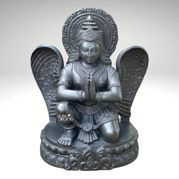 A centered eye-level studio photograph presents a meticulously hand-carved black stone statue of Garuda standing approximately two feet tall against a clean light grey gradient background. The statue is a unified piece of dark polished granite showcasing the natural finely crystalline texture of the stone. Garuda the divine eagle-man and mount of Lord Vishnu is depicted in a kneeling reverential posture balancing gracefully on his right leg while his left foot rests flat on an intricately carved circular lotus petal base. His hands are pressed together at his chest in the Anjali Mudra conveying deep devotion and respect. His serene human-like face gazes forward with a composed expression framed by elaborate traditional jewelry necklaces armlet wristlets and a tall ornate crown that is further backed by a decorative radiating prabhavali featuring a kirtimukha motif at its apex. Garuda’s iconic powerful wings are fully unfurled and spread symmetrically behind his torso filling the upper background and exhibiting detailed feather carving that contrasts with the smooth dark surface of his body. He wears a sculpted dhoti with rich patterns and a central flowing sash, all defined by precise lines. The entire sculpture demonstrates exceptional craftsmanship with sharp clean edges and smooth curves that give the dark granite a fluid, lifelike quality. The single-piece construction from durable black stone enhances its sense of ancient sanctity and spiritual presence. This image serves as an ideal visual reference for an article discussing the traditional iconography and spiritual significance of a Black Stone Garuda Statue in Namaskar Pose. The detailed depiction of the dark stone the precise execution of the prayer gesture and the impressive scale of the wings are all key elements relevant to the article's content. The two-foot height makes it a perfect size for home shrines and temple niches representing devotion and protection. The high-quality finish and accurate proportions make this particular statue an excellent representation of a traditional Hindu devotional murti.
