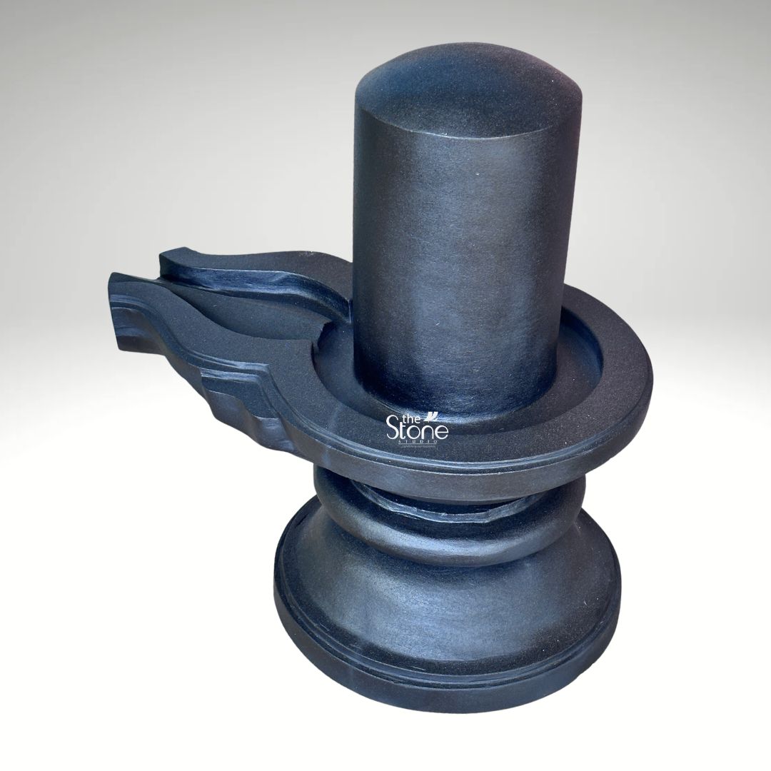 A three-quarter studio photograph captures a traditional Blackstone Shivling statue meticulously carved from a single piece of dark rich blackstone. The murti rests on a simple, rounded pedestal base which provides a stable foundation. Rising from the base is the cylindrical yoni pedestal featuring a perfectly centered smooth shivling shaft. To the side the intricately carved pranala or jaladhari extends outwards showing precise channels and traditional decorative motifs that are characteristic of fine Indian stonemasonry. The entire form has a smooth tactile quality with the dark stone absorbing and reflecting light in a way that emphasizes its heavy solid presence. This black stone Mahadev Shivling embodies the sacred union of Shiva and Shakti serving as a central object of worship in Hinduism. The precise carving of the yoni and the smooth cylindrical linga reflect traditional iconography designed for ritual bathing with water milk and other offerings. The dark solemn color of the stone evokes the infinite and formless nature of Lord Shiva making it a powerful focal point for devotion. This murti is an exemplary representation of the spiritual and aesthetic qualities that devotees seek in a Shivling for home or temple shrines. The image showcases the specific details and craftsmanship of this Blackstone Shivling statue highlighting its suitability for significant worship spaces. The clean neutral background ensures that the viewer's attention is entirely on the sculpture's form texture and traditional iconography. The photograph effectively communicates the quality of the stone and the artisan's skill providing a valuable visual reference for individuals and temple committees interested in acquiring a traditionally crafted Mahadev Shivling.