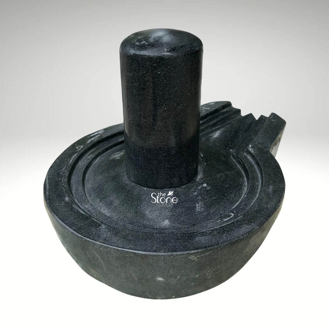 A centered eye-level studio photograph presents a beautifully hand-carved black stone Shiva Lingam with its associated Yoni base set against a clean light grey gradient background. The complete murti is carved from a single block of dense dark black granite showcasing the stone's natural finely crystalline texture and deep uniform color. The central element is the cylindrical Shiva Lingam representing the formless aspect of Lord Shiva which rises smoothly and vertically from the center of the circular Yoni pedestal. The Lingam's surface is highly polished creating a subtle sheen that reflects ambient light and contrasts with the matte finish of the surrounding elements. The Yoni base symbolizing the feminine creative energy of Goddess Parvati is a circular basin-like structure surrounding the base of the Lingam. It features a clearly defined raised circular rim and a well-proportioned projecting spout to the right side designed to channel ceremonial offerings of water or milk away from the deity. The entire Yoni base is meticulously carved from the same dark black stone, with precise concentric rings enhancing its form and balance. The carving exhibits traditional craftsmanship with smooth flowing curves and precise edges that give the Shivling and Yoni a harmonious and integrated appearance conveying a sense of ancient sanctity. This image serves as a prime visual reference for an article discussing the traditional iconography and spiritual significance of a Black Stone Shiva Lingam with Yoni. The detailed depiction of the dark granite the specific shape of the Yoni and the presence of the spout are all key elements relevant to the article's content. The single-piece construction makes it a sought-after item for home shrines and temples representing the union of masculine and feminine energies in Hindu philosophy. The high-quality finish and accurate proportions make this particular statue an excellent representation of a traditional devotional murti.