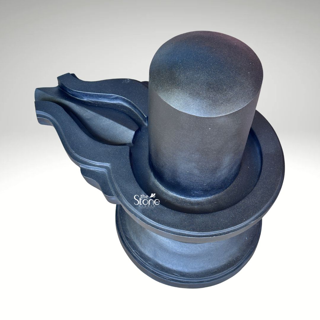 A high-angle studio photograph presents a traditional Blackstone Shivling statue meticulously carved from a single piece of dar rich blackstone. The entire murti rests on a simple rounded pedestal base providing a stable foundation. Rising from this base is the yoni pedestal featuring a smooth perfectly centered shivling shaft. The dark polished surface of the stone absorbs and reflects light subtly emphasizing the heavy solid presence of the murti and the tactile quality of the material. The most defining feature of this murti and the focus of the article is the intricately carved jaladhari. This element extends outwards from the yoni pedestal in a graceful curve designed to catch and channel the ritual offerings of water milk and other liquids during worship. The jaladhari shows precise clean channels and traditional decorative motifs a testament to the fine Indian stonemasonry involved in its creation. The smooth cylindrical form of the linga sits harmoniously within the yoni base representing the sacred union of Shiva and Shakti. This black stone Shivling with jaladhari is an exemplary representation of traditional Hindu iconography designed for ritual bathing in a home shrine or temple. The precise craftsmanship ensures that the murti is not only a spiritual focal point but also a work of art. The dark solemn color of the stone evokes the infinite and formless nature of Lord Shiva making it a powerful object for devotion. The high-quality photograph effectively communicates the rich texture of the stone and the artisan's skill providing a valuable visual reference for devotees and collectors interested in acquiring a traditionally crafted Mahadev Shivling.