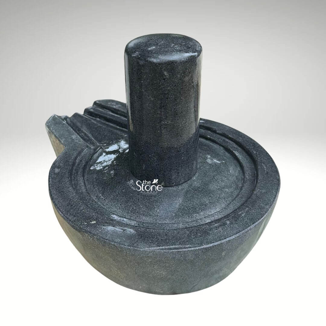 A centered eye-level studio photograph presents a meticulously hand-carved black stone Shivling with its associated Yoni base set against a clean light grey gradient background. The artifact is a single piece of dark polished black granite showcasing the natural crystalline texture and deep rich color of the stone. The central element is the cylindrical Shivling representing the formless aspect of Lord Shiva which rises smoothly and vertically from the circular Yoni pedestal. The top of the Shivling is gently domed and its surface is highly polished reflecting ambient light. The Yoni base symbolizing the feminine creative energy of Goddess Parvati is a circular basin-like structure surrounding the base of the Shivling. It features a clearly defined raised circular rim and a well-proportioned projecting spout to the left side designed to channel ceremonial offerings of water or milk away from the deity. The entire Yoni base is also crafted from the same dark black stone with subtle concentric carved rings enhancing its form. The carving exhibits traditional craftsmanship with precise lines and smooth curves that give the Shivling and Yoni a balanced and harmonious appearance. The stone has a dense durable quality conveying a sense of ancient sanctity and timeless devotion. This image serves as a prime visual reference for an article discussing the traditional iconography and spiritual significance of a Black Stone Shivling with a Yoni Base. The detailed depiction of the dark stone the specific shape of the Yoni and the presence of the spout are all key elements relevant to the article's content. The single-piece construction from durable black granite makes it a sought-after item for home shrines and temples. The high-quality finish and accurate proportions of this particular statue make it an excellent representation of a traditional Hindu devotional murti.