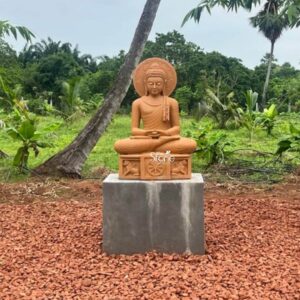 A full-view photograph captures a magnificent 4-foot tall sandstone Buddha statue standing as a prominent spiritual landmark within a lush natural garden environment in Rajahmundry Andhra Pradesh. The statue is carved from warm light-brown sandstone showcasing a natural slightly weathered texture. It is seated in the traditional vajra lotus position embodying profound peace and deep meditation. The Buddha is draped in intricately carved monastic robes with the fabric folds and a decorative patterned border clearly defined on the left shoulder. His hands are resting in his lap in the Dhyana Mudra and his face is serene, with eyes half-closed in contemplation and a gentle compassionate smile. A large intricately carved prabhavali with traditional floral motifs forms a backdrop behind his head. The statue sits upon a substantial carved rectangular pedestal that is an integral part of the entire 4-foot structure. This plinth is adorned with detailed bas-relief carvings on its front face. In the center a prominent Dharma Chakra is flanked by sculpted lions ancient symbols of the Buddha's royalty and the spread of his teachings. The entire composition is elevated on a solid simple unadorned grey concrete block creating a strong base that grounds the monumental figure. The statue's warm sandstone color provides a beautiful contrast against the surrounding natural elements. This Buddha Statue in Rajahmundry is an exceptional piece of spiritual art that harmonizes with its tropical garden setting in Andhra Pradesh. The 4-foot height ensures it is an imposing and inspiring presence visible amidst the greenery. The durable sandstone material is well-suited for the outdoor environment and weathers gracefully over time enhancing its ancient and serene appearance. The intricate carvings from the curls of the ushnisha to the figures on the base demonstrate a high level of craftsmanship making it not just a religious icon but a valuable cultural artifact. The statue is positioned amidst various plants including palm trees and other tropical foliage with the ground covered in reddish-brown gravel. The natural daylight highlights every detail of the carving creating a tranquil and reverential atmosphere perfect for quiet reflection and meditation.
