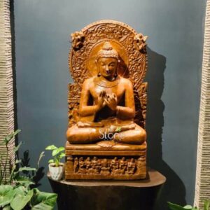A three-quarter view studio photograph captures a meticulously hand-carved three-foot-tall sandstone statue of Lord Buddha presented against a dark grey textured wall. The statue is a single piece of warm-toned, honey-colored sandstone showcasing the natural porous texture and rich grain of the stone. Lord Buddha is depicted in a seated meditative posture upon a two-tiered plinth. The front of the upper plinth is adorned with a detailed bas-relief carving featuring five small reverent figures possibly monks or devotees with offerings. The Buddha himself sits with legs crossed hands held at chest level in the dharmachakra mudra symbolizing the turning of the wheel of truth. Behind the seated Buddha an elaborate ornate prabhavali rises richly carved with intricate foliate patterns stylized figures and a prominent kirtimukha at the apex. The Buddha’s face is serene and compassionate with gently closed eye a subtle smile and a well-defined ushnisha and tightly curled hair. His body is draped in finely detailed monastic robes the folds of which are sculpted with precision, conveying a sense of naturalism despite the solid stone medium. The entire carving exhibits high-quality craftsmanship with smooth flowing curves and precise edges that give the statue a balanced and harmonious appearance. This image serves as a prime visual reference for an article discussing the presence significance, and artistry of Buddha Stone Statues in Hubli Karnataka. The specific details such as the warm sandstone material the three-foot height the intricate prabhavali and the dharmachakra mudra are crucial for those seeking authentic devotional art in the region. The statue's aesthetic blends traditional Indian temple carving styles with the specific iconography of Buddhism making it an ideal centerpiece for any sacred or contemplative space. The warm honey-colored stone makes it a sought-after choice for home shrines and public spaces alike and this visual representation captures its texture proportion and serene presence perfectly.
