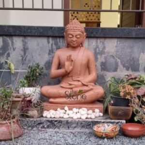 A centered eye-level photograph captures a reverently installed two-foot-tall sandstone Buddha statue positioned outdoors against a rustic textured natural stone wall that shows signs of weathering. The statue is the central element seated in a meditative lotus posture upon a low grey stone plinth. The sandstone exhibits a warm earthy pinkish-brown hue and a matte porous texture typical of hand-chiseled stone. Lord Buddha is depicted with a serene compassionate expression gently closed eyes and tightly curled hair in the traditional ushnisha. His right hand is raised at chest level in the vitarka mudra with the thumb and index finger touching while his left hand rests in his lap in the dhyana mudra. His body is draped in finely detailed monastic robes the folds of which are sculpted with precision conveying a sense of natural flowing fabric despite the solid medium. The base of the statue is adorned with a collection of smooth white river pebbles creating a clean naturalistic border on the plinth. The statue is surrounded by a variety of potted plants and garden elements arranged as a personal shrine. To the lower left a rustic weathered terracotta pot and various trailing plants with purple and green leaves are visible. To the lower right a cluster of diverse potted succulents and a leafy plant in a brown square ceramic pot add greenery. On the ground in front of the plinth two decorative bowls rest: one small colorful patterned bowl and a plain terracotta bowl. The background shows a glimpse of a modern building with clean lines and a decorative railing creating an interesting contrast with the traditional art and natural garden setting. This image serves as a prime visual reference for an article discussing the cultural significance and installation of Buddha stone statues in Kolhapur. The specific details such as the warm sandstone material the two-foot height the detailed mudra and the natural outdoor installation with traditional elements like terracotta and pebbles are crucial for those seeking authentic devotional art in the region. The setting demonstrates how such a statue can be reverently enshrined making the image highly relevant for articles discussing the selection placement and decoration ideas for Buddha statues in Kolhapur homes and gardens. The craftsmanship of the sandstone carving is evident making the statue an attractive centerpiece for any sacred space.