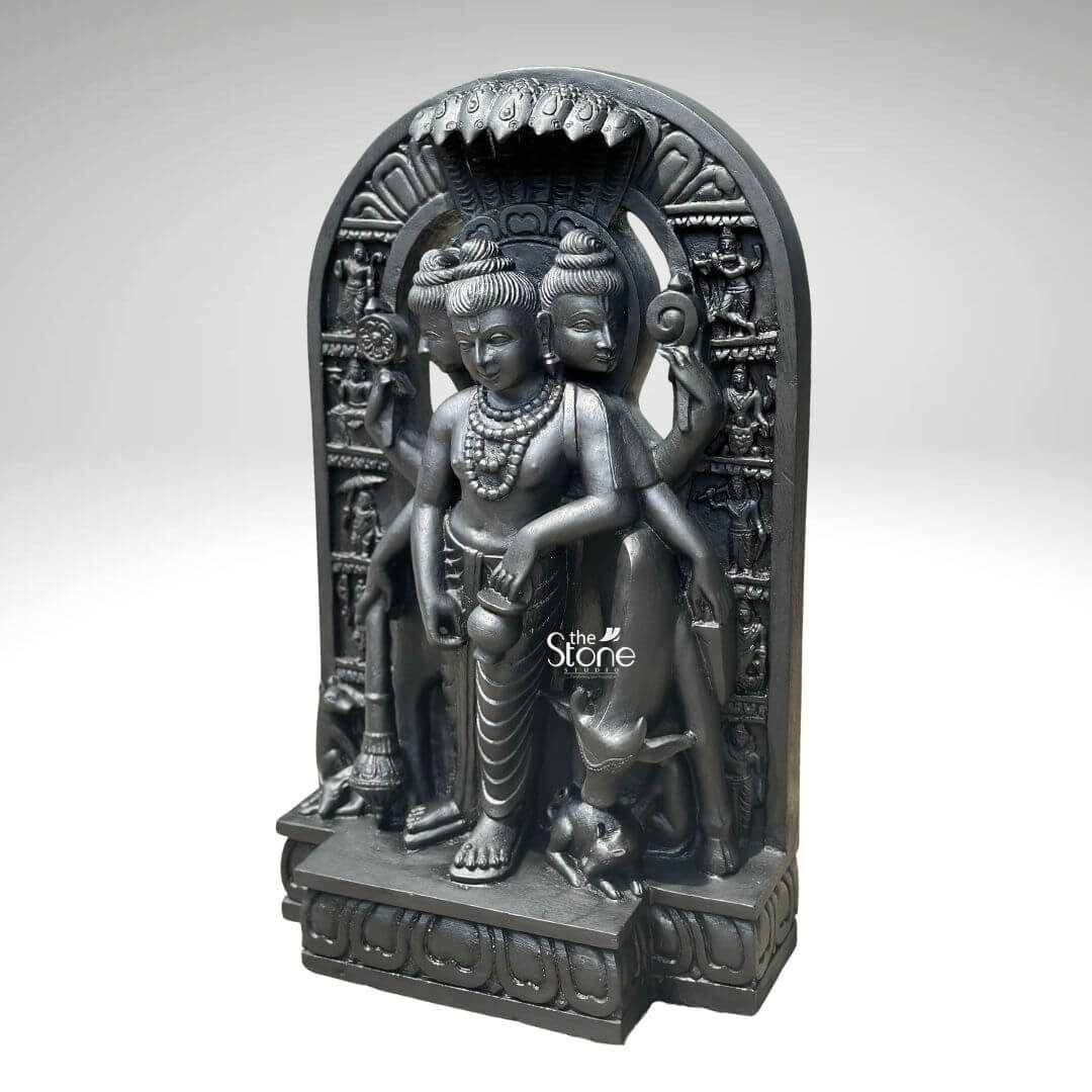 This studio photograph presents a finely detailed 2-foot tall statue of Datta Guru also known as Dattatreya carved from a single block of rich dark black natural stone. The statue depicts the Hindu deity in a standing posture embodying the combined form of the Trimurti: Brahma Vishnu and Shiva. Dattatreya is shown with three distinct faces each serene and adorned with traditional crowns and elaborate jewelry symbolizing the three-in-one nature of the god. The smooth polished surface of the blackstone highlights the intricate craftsmanship and the semi-precious quality of the material making it a perfect Datta Guru Stone Statue for worship or home decor. The deity is depicted with six arm holding symbolic attributes associated with the three gods including a trident conch shell discus water pot and a mala. He is clad in simple dhotis and adorned with numerous strands of rudraksha beads and gemstone necklaces all rendered with precision in the black stone. The central form is flanked by his four canine companions representing the four Vedas and a cow stands behind him completing the traditional iconography. The statue stands on an ornate multi-tiered lotus pedestal adding to its divine presence. The entire sculpture is set within a carved arched niche that is further embellished with smaller relief carvings of various sages and disciples. The dark black stone creates a striking contrast against the clean neutral grey background which is free of any distractions. The lighting is focused and directional casting soft shadows that emphasize the three-dimensional form and the deep relief work on the crown jewelry and base making this 2ft Dattatreya Black stone statue a beautiful and spiritually significant piece.