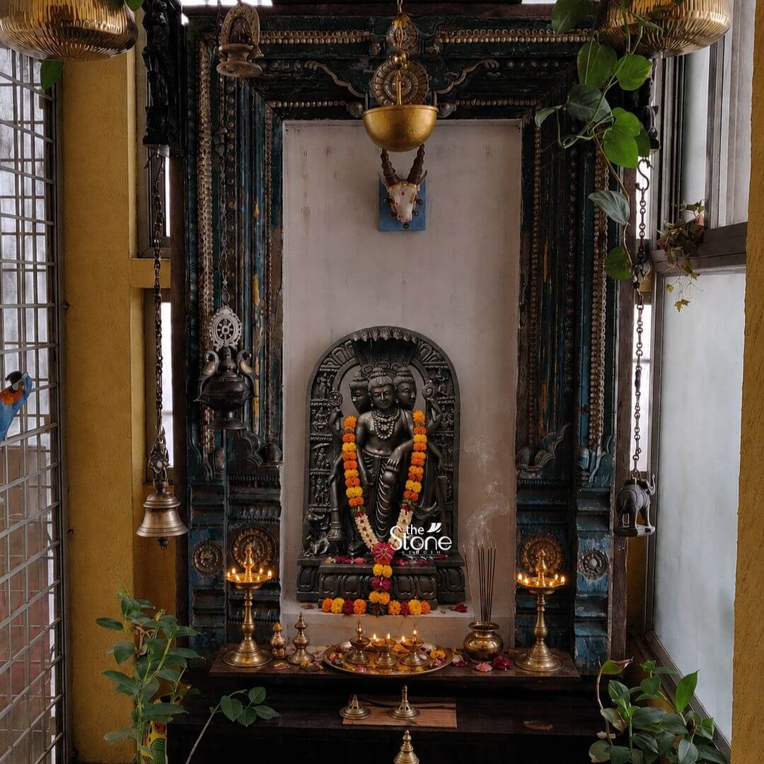 This finely crafted 2-foot tall black stone statue of Lord Dattatreya is presented within a traditional home shrine showcasing the complete iconography described in the article Dattatreya Idol with Cow and Dogs. The deity carved from a single block of deep black granite is depicted in a standing posture embodying the combined form of the Trimurti Brahma Vishnu and Shiva. He has three distinct serene faces each adorned with detailed crowns multiple strands of rudraksha beads and intricate necklaces. The statue exhibits remarkable sculptural depth and texture with six arms holding symbolic attributes such as a trident conch shell discus water pot and mala. The four canine companions representing the four Vedas are carefully carved at his feet while the divine cow Kamadhenu stands attentively behind him looking over his shoulder. The entire murti is draped in a vibrant multi-layered garland of yellow and orange marigold flowers with a central strand of pearls and colorful beads adding a traditional touch of worship. The statue is housed within a richly carved antique dark wood frame that serves as a decorative arch for the shrine. The shrine itself is set upon a dark wood altar covered with a red cloth and scattered marigold flowers. The surrounding environment enhances the sacred atmosphere featuring two lit brass oil lamps and a small ornate brass pot holding incense sticks from which a gentle wisp of smoke rises. Various other traditional brass puja items including small pots and a conch are arranged on the altar making it a beautiful representation of a Dattatreya Idol with Cow and Dogs in a home temple setting.