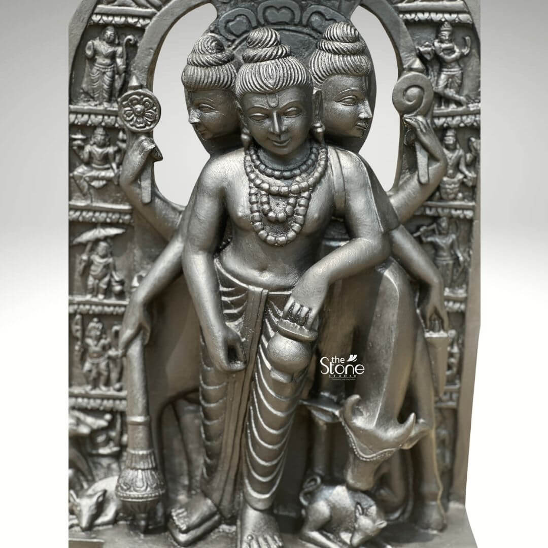 This studio photograph presents a finely detailed 2-foot tall statue of Lord Dattatreya, meticulously hand-carved from a single block of rich, dark green natural stone. The statue depicts the Hindu deity in a standing posture known as the combined form of the Trimurti Brahma Vishnu and Shiva. The deity is shown with three distinct serene faces each adorned with carved crowns multiple strands of rudraksha beads and intricate necklaces symbolizing the three-in-one nature of the god. The smooth yet textured surface of the blackstone highlights the skilled artisanship and traditional iconography making it a perfect Dattatreya Murti in Black Stone for worship or home decor. The idol exhibits remarkable sculptural depth and detail. Dattatreya is depicted with six arms holding symbolic attributes associated with the three gods including a trident conch shell discus water pot and a mala. He is clad in simple dhotis and adorned with numerous strands of beads and garlands all rendered with precision in the dark green stone. The central form is flanked by his four canine companions representing the four Vedas carefully carved at his feet while the divine cow Kamadhenu stands attentively behind him looking over his shoulder. The entire sculpture is framed by a deeply carved archway further embellished with smaller relief carvings of various sages and disciples. The dark green stone creates a striking contrast against the clean neutral grey background which is free of any distractions. The lighting is focused and directional casting soft shadows that emphasize the three-dimensional form and the intricate relief work on the crowns jewelry and base making this image an excellent representation of a quality Dattatreya Murti in Black Stone.