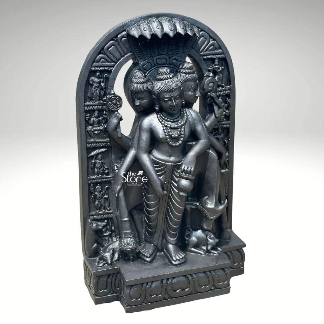 This studio photograph presents a finely detailed 2-foot tall statue of Lord Dattatreya Swamy, meticulously carved from a single block of rich dark black natural stone. The statue depicts the Hindu deity in a standing posture known as the combined form of the Trimurti Brahma Vishnu and Shiva. The deity is shown with three distinct, serene faces each adorned with carved crowns multiple strands of rudraksha beads and intricate necklaces symbolizing the three-in-one nature of the god. The smooth yet textured surface of the blackstone highlights the skilled artisanship and traditional iconography making it a perfect Dattatreya Swamy Idol for Temple worship or home shrines. The idol exhibits remarkable sculptural depth and detail. Dattatreya is depicted with six arms holding symbolic attributes associated with the three gods including a trident conch shell discus water pot and a mala. He is clad in simple dhotis and adorned with numerous strands of beads and garlands all rendered with precision in the black stone. The central form is flanked by his four canine companions representing the four Vedas carefully carved at his feet while the divine cow Kamadhenu stands attentively behind him looking over his shoulder. The entire sculpture is framed by a deeply carved archway further embellished with smaller relief carvings of various sages and disciples. The dark black stone creates a striking contrast against the clean neutral grey background which is free of any distractions. The lighting is focused and directional casting soft shadows that emphasize the three-dimensional form and the intricate relief work on the crowns jewelry and base making this image an excellent representation of a quality Dattatreya Swamy Idol for Temple.