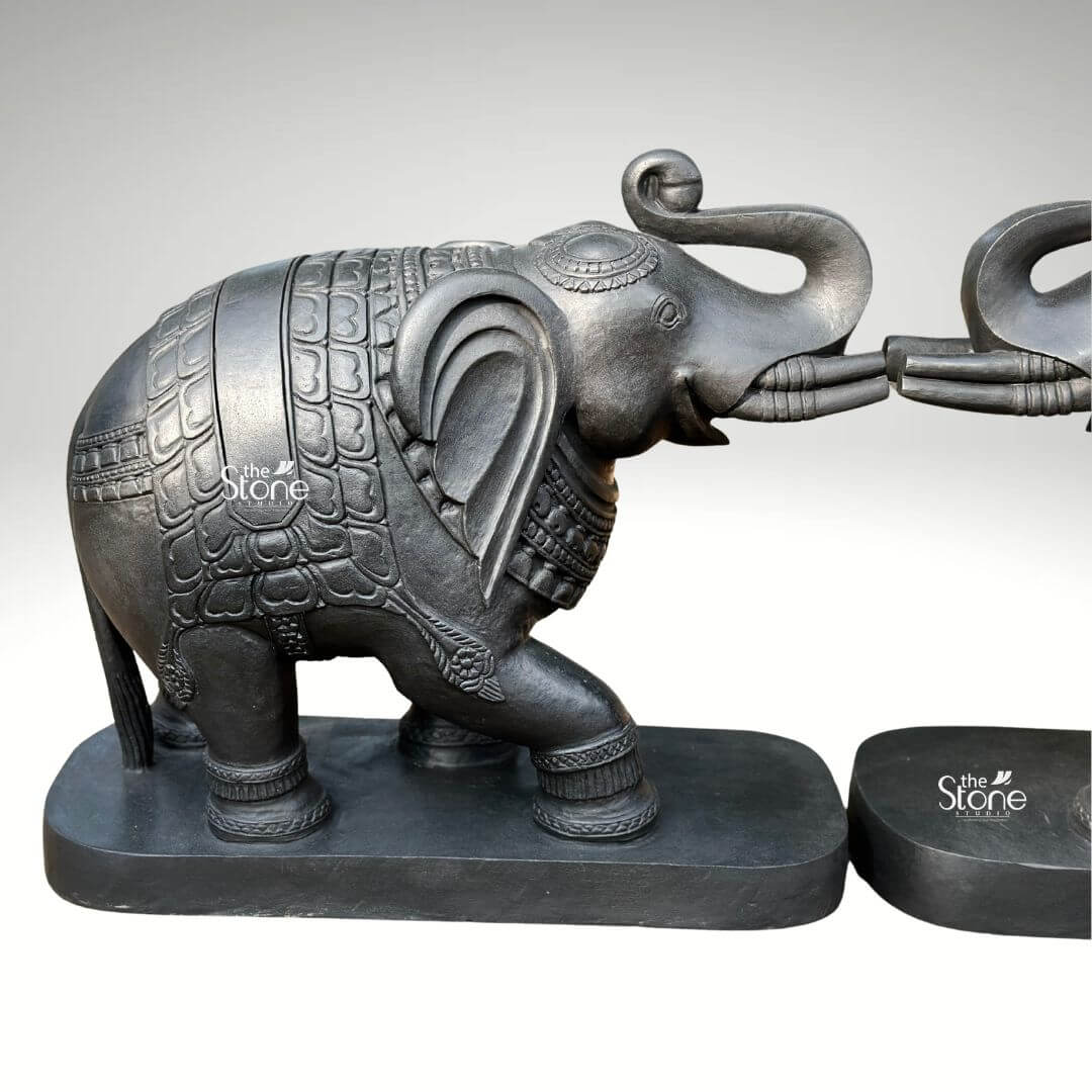A close-up three-quarter view photograph captures a magnificent pair of 26-inch blackstone elephant statues meticulously hand-carved and arranged with their trunks touching in a gesture of affection and greeting, suitable for a main entrance. The elephants are crafted from dark rich blackstone, giving them a smooth, polished texture that highlights the intricate details of their form. Each elephant is adorned with finely etched patterns on their backs resembling ornate decorative blankets and has detailed carvings on their legs ears and trunks showcasing a high level of traditional Indian craftsmanship. The elephants are depicted with their trunks curled and interlocked a pose symbolizing good luck strength and prosperity common in Indian elephant iconography. The pair of statues is positioned symmetrically on a simple unadorned blackstone base which grounds the monumental figures. The elephants' substantial 26-inch length makes them an imposing and inspiring presence suitable for a grand home entrance consistent with the article's theme of a decorative elephant pair for main entrance. The dark stone material provides a powerful and elegant contrast against a clean neutral light-grey background allowing every nuance of the carving to be appreciated. Even directional studio lighting illuminates the pair emphasizing the deep textures and sharp lines of the carvings across the dark polished surface of the stone. This image is an exceptional representation of a traditional blackstone elephant statue pair showcasing the rich cultural artistry and spiritual significance of such a piece destined to grace a home's main entrance. The fine details from the delicate patterns on the decorative blankets to the lifelike form of the raised and touching trunks and flowing tails demonstrate the artisan's skill in working with this durable and classic medium. The statues' posture with the elephants facing each other with touching trunks conveys a deep sense of reverence strength and good fortune creating a welcoming and spiritually balanced atmosphere for all who enter. The clean presentation allows the viewer to appreciate the full form and intricate carvings of the 26-inch blackstone murtis.