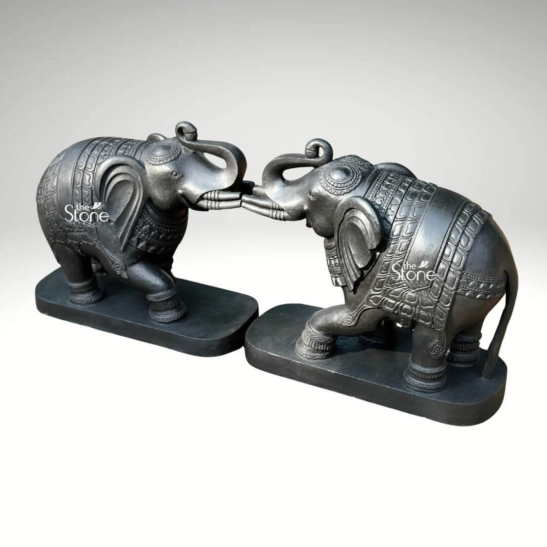 A detailed three-quarter view photograph captures a magnificent pair of 26-inch blackstone elephant statues meticulously hand-carved and arranged with their raised trunks touching in a gesture of affection and greeting. The elephants are crafted from dark rich blackstone giving them a smooth, polished texture that highlights the intricate details of their form. Each elephant is adorned with finely etched patterns on their backs resembling ornate decorative blankets and has detailed carvings on their legs ears and trunks showcasing a high level of traditional Indian craftsmanship. The elephants are depicted with their raised trunks curled and interlocked a pose symbolizing good luck strength and prosperity common in Indian elephant iconography. The pair of statues is positioned symmetrically on simple unadorned blackstone bases which ground the monumental figures. The elephants' substantial 26-inch length makes them an imposing and inspiring presence suitable for a grand home interior entryway or living space consistent with the article's theme of a Decorative Elephant Statue Pair with Raised Trunks. The dark stone material provides a powerful and elegant contrast against a clean neutral light-grey background allowing every nuance of the carving to be appreciated. Even directional studio lighing illuminates the pair emphasizing the deep textures and sharp lines of the carvings across the dark polished surface of the stone. This image is an exceptional representation of a traditional blackstone elephant statue pair showcasing the rich cultural artistry and spiritual significance of such a piece. The fine details from the delicate patterns on the decorative blankets to the lifelike form of the raised and touching trunks and flowing tails demonstrate the artisan's skill in working with this durable and classic medium. The statues' posture with the elephants facing each other with touching raised trunks, conveys a deep sense of reverence strength and good fortune suitable for creating a welcoming and spiritually balanced atmosphere. The clean presentation allows the viewer to appreciate the full form and intricate carvings of the 26-inch blackstone murtis.