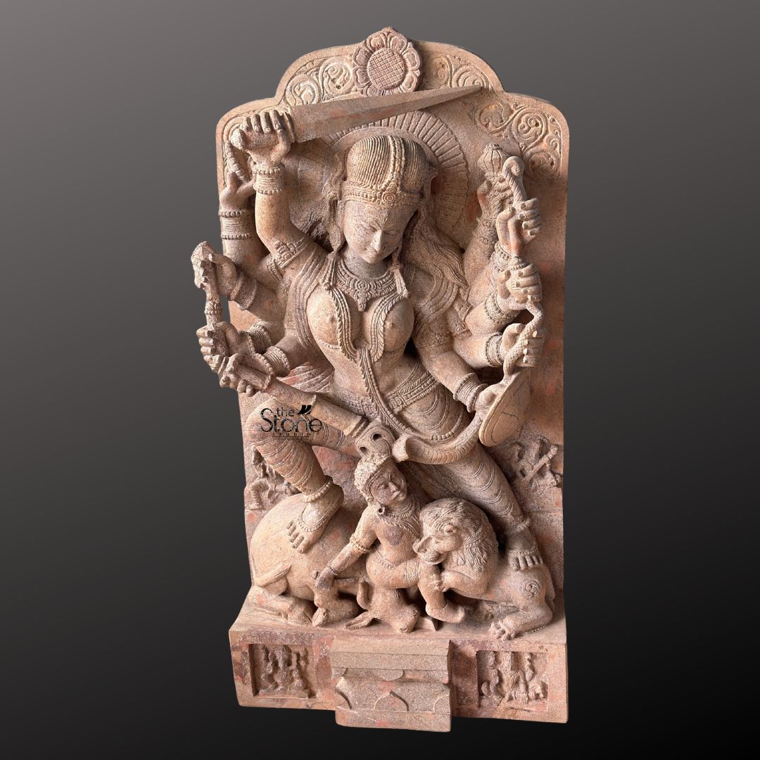 This magnificent stone sculpture carved from a warm-toned pinkish-red sandstone, depicts the Hindu goddess Durga in her powerful form as Mahishasura Mardini the slayer of the buffalo demon. The statue is a high-relief stele showcasing intricate classical Indian artistry. Durga is shown in a dynamic active posture, mid-battle, with her right foot planted firmly on the severed head of the buffalo demon and her left leg bent. Her primary right hand wields a sword held aloft while her primary left hand holds the tail of the demon who is emerging in human form from the buffalo's carcass. The goddess is depicted with eight arms each holding a symbolic weapon or item including a trident conch disc bow and arrow all rendered in fine detail. She wears elaborate jewelry a detailed crown and a flowing dhoti. The demon Mahishasura is shown in a contorted defeated posture beneath her foot with a lion mount attacking him from the side. The entire sculpture is framed by an ornate archway with floral and geometric carvings, and the base includes small relief figures and a central plinth emphasizing the sacred nature of this Mahishasura Mardini Durga statue in Sandstone. The statue is presented against a dark neutral grey background which allows the natural texture and warm color of the sandstone to stand out. The lighting is directional casting soft shadows that enhance the depth and three-dimensionality of the complex carving, showcasing the skilled artisanship. This image is an ideal representation for an article discussing the iconography spiritual significance and cultural heritage of the Durga Devi Mahishasura Mardini Statue.