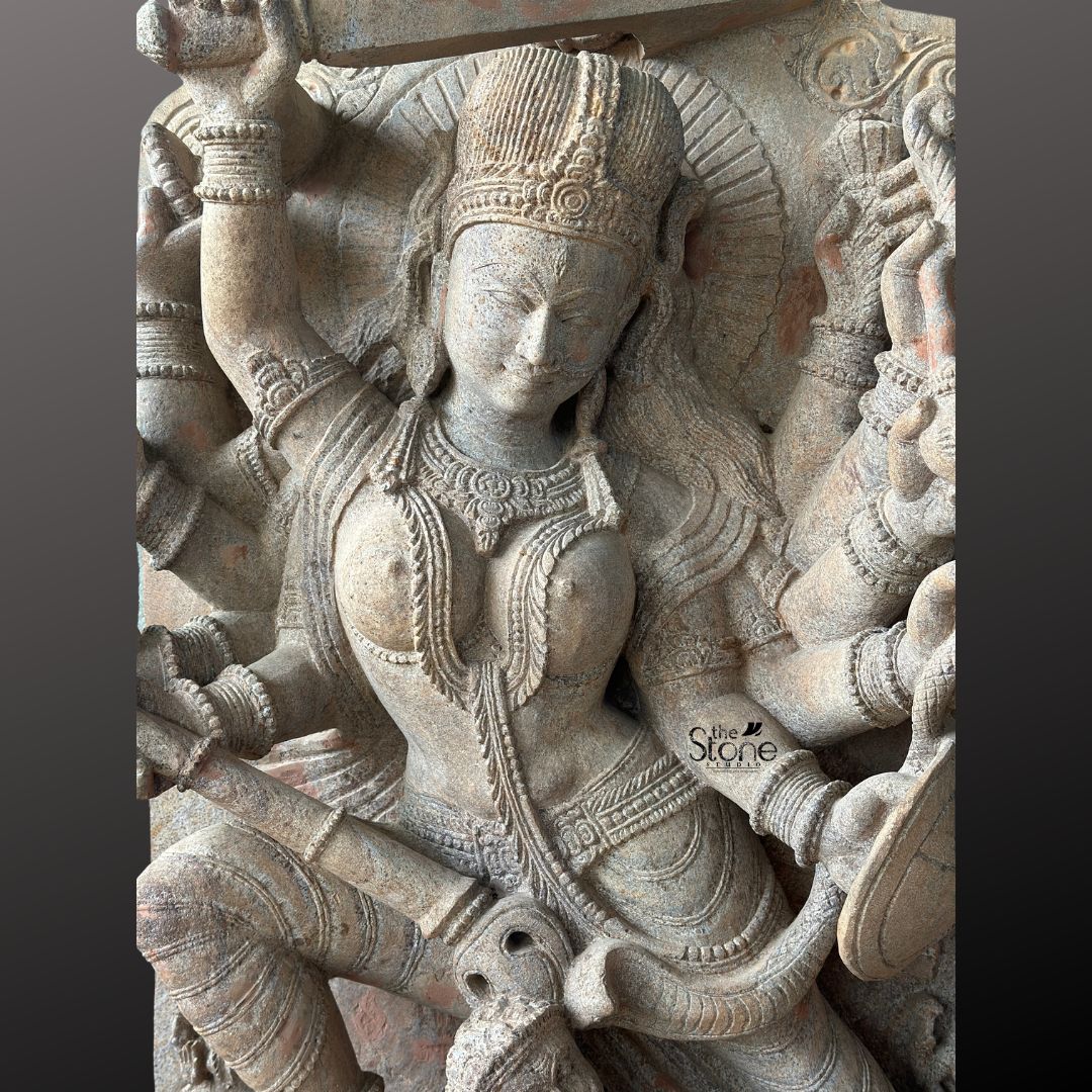 This close-up studio photograph captures a richly detailed high-relief sandstone carving of the Hindu goddess Durga in her fierce aspect as Mahishasura Mardini the slayer of the buffalo demon. The statue crafted from a warm mottled beige sandstone showcases the intricate skill of traditional Indian stone artistry. The central figure is the multi-armed Durga depicted in a dynamic powerful posture mid-battle. Her face framed by a detailed crown and flowing hair holds a serene yet focused expression as she vanquishes the demon. She wears elaborate jewelry armlets and a flowing dhoti all rendered with fine deep-cut detail that emphasizes the three-dimensional form. Durga is shown with numerous arms each holding a symbolic weapon or item. Her primary right hand wields a sword held aloft behind her head while another right hand holds a trident piercing the demon below. Her primary left hand holds the tail of the demon who is emerging from the carcass of a buffalo. The carving clearly displays the buffalo demon Mahishasura in a contorted defeated posture beneath her main foot. A lion mount is also visible attacking the demon from the side. The statue is set against a dark neutral grey background which allows the natural texture and warm color of the sandstone to stand out. The lighting is directional casting soft shadows that enhance the depth and three-dimensionality of the complex carving making the scene come alive. This image is an ideal representation for an article discussing the iconography spiritual significance and cultural heritage of a Durga Maa Stone Statue with Mahishasura.
