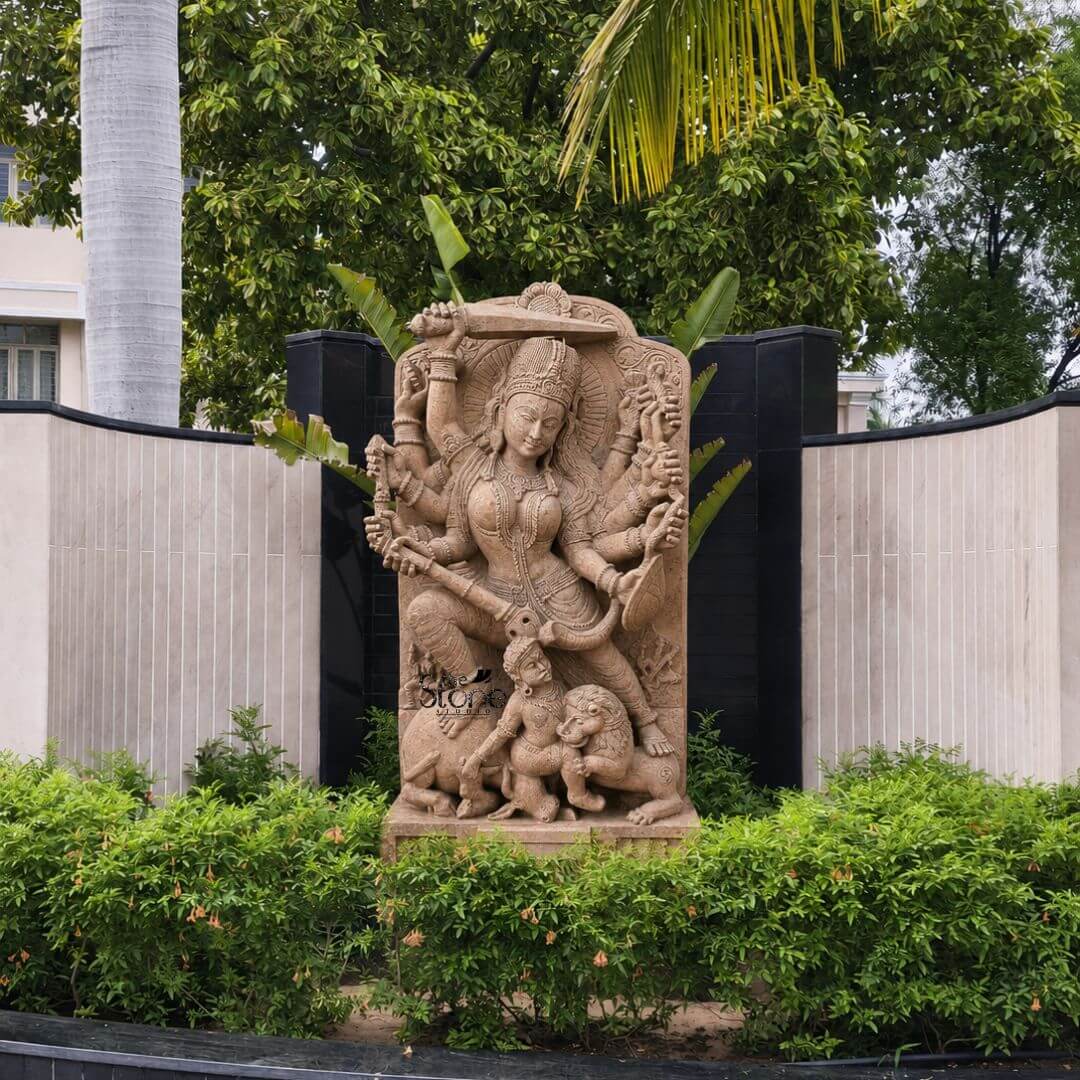 This magnificent stone sculpture carved from warm-toned sandstone depicts the Hindu goddess Durga in her powerful form as Mahishasura Mardini the slayer of the buffalo demon. The statue is a high-relief stele showcasing intricate classical Indian artistry. Durga is shown in a dynamic active posture mid-battle with her right foot planted firmly on the severed head of the buffalo demon and her left leg bent. Her primary right hand wields a sword held aloft while her primary left hand holds the tail of the demon who is emerging in human form from the buffalo's carcass. The goddess is depicted with eight arms each holding a symbolic weapon or item including a trident conch disc bow and arrow all rendered in fine detail. She wears elaborate jewelry a detailed crown and a flowing dhoti. The demon Mahishasura is shown in a contorted defeated posture beneath her foot with a lion mount attacking him from the side. The entire sculpture is framed by an ornate archway with floral and geometric carvings, and the base includes small relief figures and a central plinth emphasizing the sacred nature of this Mahishasura Mardini Durga statue in Sandstone. The statue is installed outdoors as a focal point within a landscaped garden set against a curved wall with alternating light stone and dark panels flanked by lush green hedges and mature trees including a prominent palm. This placement illustrates its suitability as a Durga Mata Stone Idol for Temple or shrine environments creating a serene and spiritual atmosphere. Natural daylight illuminates the sculpture casting soft shadows that enhance the depth and three-dimensionality of the complex carving, showcasing the skilled artisanship and the rich texture of the sandstone.