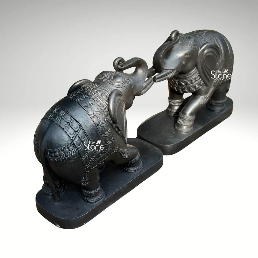 A high-angle three-quarter view studio photograph captures a magnificent pair of 26-inch blackstone elephant statues meticulously hand-carved and positioned to face each other. The elephants are arranged in an affectionate pose with their trunks curled upward and interlocked a traditional gesture symbolizing greeting strength and good fortune. They are crafted from a dark rich blackstone giving the entire sculpture a smooth polished texture that highlights the deep charcoal-like color and subtle natural variations of the stone. Both figures are mounted on matching unadorned black rectangular stone bases which ground the monumental figures and provide a stable foundation. The artistry of this blackstone elephant statue pair is evident in the fine intricate carvings that adorn their forms. Each elephant is embellished with detailed etched patterns covering their backs resembling an ornate decorative blanket as well as on their ears headpieces and legs. The level of traditional craftsmanship is exceptional with sharp clean lines and deep textures that catch the light showcasing the artisan's skill in rendering lifelike forms from a durable medium. From the delicate scale-like patterns on the blankets to the lifelike wrinkles on their trunks and the gentle curve of their tusks every detail contributes to a sense of grandeur and elegance. Designed as a striking centerpiece this blackstone elephant statue pair for home entrance is perfectly scaled to create a commanding yet welcoming presence. The dark elegant stone material provides a powerful contrast against a clean neutral light-grey background allowing every nuance of the carving to be appreciated. The symmetrical arrangement and touching trunks convey a deep sense of harmony and balance making this sculpture ideal for flanking a grand entryway or decorating a living space. This image effectively communicates the rich cultural artistry imposing size and spiritual significance of such a decorative piece reinforcing its role in creating an auspicious and spiritually balanced atmosphere.