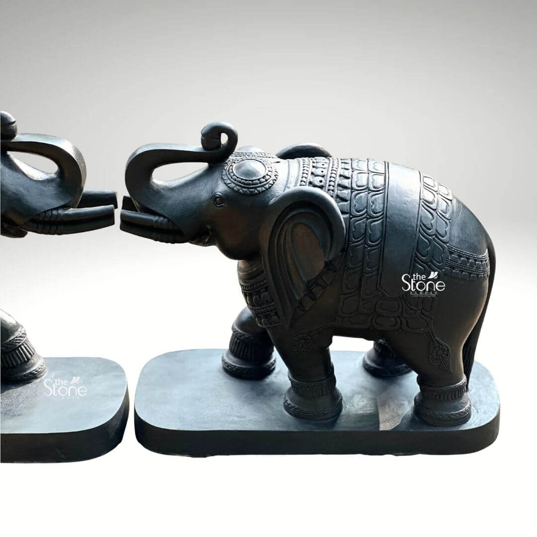 A detailed studio product photograph captures a magnificent pair of 26-inch blackstone elephant statues presented against a neutral light-grey background. The two statues are arranged symmetrically facing each other with their raised trunks gently touching a traditional pose symbolizing affection greeting and good fortune. The statues are meticulously hand-carved from dark rich blackstone which has been polished to a smooth matte finish that accentuates the deep color and subtle natural grain of the stone. Each figure is mounted on a simple unadorned black rectangular stone base which grounds the monumental figures and provides stability. The artistry and craftsmanship of this blackstone elephant pair are showcased through intricate fine carvings that adorn their forms. The surface of each elephant is covered in detailed etched patterns resembling an ornate decorative blanket complete with traditional Indian motifs, scale-like textures and geometric borders. The level of skill is further evident in the carved details of the ears, headpieces tusks and the expressive form of the interlocking trunks and flowing tails. The sharp, precise lines of the carvings contrast beautifully with the smooth dark stone creating a visually rich and textured surface. As a perfect example of an Elephant Sculpture Pair for Home these 26-inch blackstone murtis are designed to be a commanding yet elegant presence in an entryway or living space. The dark sophisticated material provides a powerful focal point and the symmetrical facing arrangement conveys a sense of balance and harmony. The image effectively communicates the substantial scale cultural significance and spiritual meaning of the sculpture making it an ideal decorative piece for fostering an auspicious and welcoming atmosphere in a home. The high-quality lighting ensures that every nuance of the hand-carving is visible highlighting the rich cultural artistry of the set.