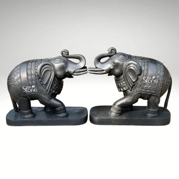 A full-view studio photograph displays a magnificent pair of 26-inch blackstone elephant statues meticulously hand-carved and arranged facing each other in a mirrored pose. The elephants are crafted from dark polished blackstone giving them a smooth rich texture that highlights the intricate details of their form. Each elephant is adorned with finely etched patterns on their backs resembling ornate decorative blankets and has detailed carvings on their legs ears and trunks showcasing a high level of traditional Indian craftsmanship. The elephants are depicted with their trunks raised in a trumpet-like gesture symbolizing good luck strength and prosperity common in Indian elephant iconography. The pair of statues is positioned symmetrically on a simple unadorned blackstone base which grounds the monumental figures. The elephants' substantial 26-inch length makes them an imposing and inspiring presence suitable for a grand home interior entryway or living space consistent with the article's theme of an Elephant Statue Pair for Home Decor. The dark stone material provides a powerful and elegant contrast against a clean neutral light-grey background, allowing every nuance of the carving to be appreciated. Even directional studio lighting illuminates the pair emphasizing the deep textures and sharp lines of the carvings across the dark polished surface of the stone. This image is an exceptional representation of a traditional Elephant Statue Pair in Blackstone showcasing the rich cultural artistry and spiritual significance of such a piece destined for home decoration. The fine details from the delicate patterns on the decorative blankets to the lifelike form of the raised trunks and flowing tails demonstrate the artisan's skill in working with this durable and classic medium. The statues' posture with the elephants facing each other with raised trunks conveys a deep sense of reverence strength and good fortune suitable for creating a welcoming and spiritually balanced atmosphere in a home. The clean presentation allows the viewer to appreciate the full form and intricate carvings of the 26-inch blackstone murtis.