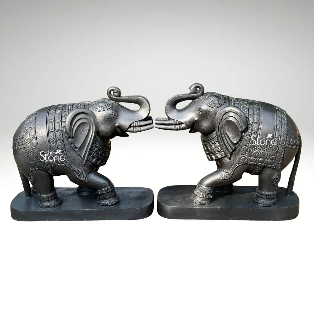 A full-view studio photograph displays a magnificent pair of 26-inch blackstone elephant statues meticulously hand-carved and arranged facing each other in a mirrored pose. The elephants are crafted from dark polished blackstone giving them a smooth rich texture that highlights the intricate details of their form. Each elephant is adorned with finely etched patterns on their backs resembling ornate decorative blankets and has detailed carvings on their legs ears and trunks showcasing a high level of traditional Indian craftsmanship. The elephants are depicted with their trunks raised in a trumpet-like gesture symbolizing good luck strength and prosperity common in Indian elephant iconography. The pair of statues is positioned symmetrically on a simple unadorned blackstone base which grounds the monumental figures. The elephants' substantial 26-inch length makes them an imposing and inspiring presence suitable for a grand home interior entryway or living space consistent with the article's theme of an Elephant Statue Pair for Home Decor. The dark stone material provides a powerful and elegant contrast against a clean neutral light-grey background, allowing every nuance of the carving to be appreciated. Even directional studio lighting illuminates the pair emphasizing the deep textures and sharp lines of the carvings across the dark polished surface of the stone. This image is an exceptional representation of a traditional Elephant Statue Pair in Blackstone showcasing the rich cultural artistry and spiritual significance of such a piece destined for home decoration. The fine details from the delicate patterns on the decorative blankets to the lifelike form of the raised trunks and flowing tails demonstrate the artisan's skill in working with this durable and classic medium. The statues' posture with the elephants facing each other with raised trunks conveys a deep sense of reverence strength and good fortune suitable for creating a welcoming and spiritually balanced atmosphere in a home. The clean presentation allows the viewer to appreciate the full form and intricate carvings of the 26-inch blackstone murtis.