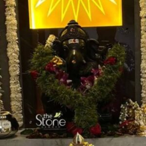 The central focus of the image is a finely detailed 18-inch black stone statue of the Hindu deity Ganesha depicted in a traditional seated posture. The statue made of polished black granite displays intricate carvings on the crown jewelry and the folds of the dhoti highlighting the craftsmanship. The Ganesha figure is draped in a lush V-shaped garland of green foliage and interspersed with deep red roses and delicate purple orchids adding a vibrant contrast to the dark stone. The statue is positioned within what appears to be a home shrine in Mumbai suggesting its suitability as a Ganesha statue for home worship. It is placed on a decorated altar with the surrounding environment including traditional elements like a silver metal kalash to the left and a small ornate lamp base at the bottom center. The vertical pillars flanking the shrine are heavily adorned with long garlands of white jasmine flowers enhancing the sacred ambiance of a Mumbai home setup. Above the statue a bright, golden-yellow backdrop is visible featuring a stylized sunburst or abstract floral pattern which illuminates the deity. This backdrop combined with the floral garlands and the dark stone of the Ganesha statue creates a focused devotional space. The lighting is warm and directional casting soft shadows that emphasize the three-dimensional form and the serene expression on Ganesha’s face making it an ideal depiction of a Ganesha statue for home settings.