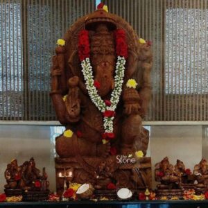 A magnificent 5-foot-tall Ganesha statue hand-carved from warm-toned sandstone stands as the majestic focal point within a beautifully decorated hotel lobby or reception area in Visakhapatnam. The monolithic murti depicts Lord Ganesha in a seated benevolent posture known as Asanasth Ganesha with four arms. He is adorned with elaborate traditional jewelry including multiple necklaces armlets and anklets all intricately sculpted from the same durable sandstone. A large ornate halo with traditional patterns frames his head adding to the divine aura. The natural porous texture and warm earthy color of the sandstone are beautifully highlighted by the warm ambient lighting of the hotel space giving the sculpture a rich tactile quality. The massive 5-foot Ganesha statue is elegantly arrayed for daily Puja making it an inviting and spiritually resonant centerpiece for guests arriving at the hotel in Visakhapatnam. The statue is draped in long fresh garlands of white jasmine interspersed with vibrant red roses that cascade down his form creating a beautiful contrast against the natural stone. Additional offerings of individual red roses and yellow marigolds are scattered around the base of his pedestal. In the foreground traditional brass oil lamps cast a soft flickering glow alongside fresh coconuts and more scattered flower petals demonstrating an ongoing reverence and creating a welcoming and auspicious atmosphere. This image serves as an excellent illustration for an article about sourcing a significant Ganesha Statue for Hotel in Visakhapatnam. The photograph captures the imposing scale cultural artistry and spiritual significance that such a large sandstone murti brings to a hospitality setting. The fine details of the carving from the gentle curve of his trunk to the delicate folds of his dhoti and the decorative patterns on his crown, are clearly visible showcasing the high level of traditional stonemasonry. The statue's placement conveys a deep sense of tradition and a warm cultural welcome to all who enter the hotel.