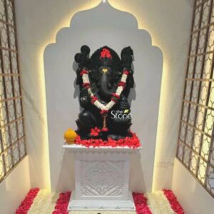 A detailed eye-level photograph captures a magnificent 3-foot Ganesha Stone Statue meticulously hand-carved from blackstone standing as the revered centerpiece within a home temple alcove. The statue depicts the deity seated in a traditional posture adorned with an intricate garland of fresh white jasmine and red rose petals. The fine details of the blackstone carving are evident in the crown jewelry and the four arms holding symbolic attributes. The statue is positioned on a white ornately carved marble pedestal which is richly covered in a scatter of loose red hibiscus flowers and green leaves with a small round golden-yellow offering placed to the left. The sacred setting is enhanced by the arched illuminated niche in the wall behind the statue featuring a warm, back-lit glow that highlights the dark form of the Ganesha Stone Statue. The alcove is flanked by decorative geometric-patterned wooden screens adding a touch of traditional Maharashtrian architectural elegance. The floor directly in front of the pedestal is meticulously decorated with a square rangoli-like border of alternating white jasmine and red rose petals framing the sacred space. The composition creates a harmonious blend of dark stone and vibrant floral colors. This image is an authentic representation of a consecrated Ganesha Stone Statue reflecting the rich cultural and spiritual traditions of Ahilyanagar Maharashtra where such blackstone artistry is highly valued. The 3-foot height of the statue makes it an imposing yet accessible presence for daily worship in a domestic setting. The overall scene conveys a sense of deep devotion and reverence making it a perfect visual for an article exploring the significance and craftsmanship of Ganesha statues from this region. The lighting is soft and focused drawing the viewer's eye directly to the divine form.