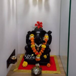 This finely crafted blackstone Ganesha statue is presented within a home altar setting showcasing the quality and traditional artistry available from sources for Ganesha Stone Statues in Thane. The deity is carved from a single block of deep black natural stone giving it a rich dark patina that contrasts beautifully with its surroundings. Ganesha is depicted in a traditional seated posture known as Lalitasana with his right leg folded and his left leg pendant resting on a matching black stone base. The statue is meticulously adorned for worship making it a prime example of a Ganesha statue in blackstone used for home shrines. He wears a vibrant multi-layered garland made of alternating yellow marigold and orange marigold flowers with a central strand of white pearls and colorful beads. A single striking red hibiscus flower is placed atop his crown adding a traditional touch of color. His forehead features a traditional white tripundra tilak and his eyes are clearly defined capturing a serene expression. The carving shows delicate details in the crown jewelry and the folds of his garment. The Ganesha idol rests on a square red velvet cloth with a decorative gold-embroidered border placed on a light wood shelf. The surrounding shrine elements enhance the spiritual atmosphere including a traditional brass oil lamp on a wall sconce to the left and a silver-toned metal pot with a decorative lid in the foreground. Loose marigold flowers are scattered on the red cloth near the base of the statue and a small cluster of white flowers sits in front of the metal pot. The background is a simple neutral wall ensuring the focus remains entirely on the blackstone Ganesha statue.