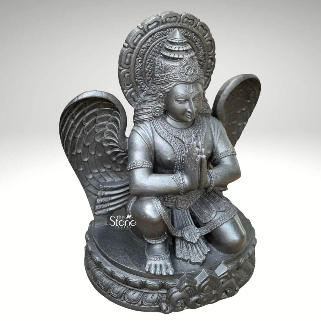 A three-quarter view studio photograph presents a meticulously hand-carved black stone statue of Garuda, standing approximately two feet tall against a clean light grey gradient background. The statue is a unified piece of dark polished granit showcasing the stone's natural finely crystalline texture and deep, uniform color. Garuda the divine eagle-man and mount of Lord Vishnu is depicted in a kneeling, reverential posture, balancing gracefully on his right leg while his left foot rests flat on an intricately carved, circular lotus petal base. His hands are pressed together at his chest in the Anjali Mudra conveying deep devotion and respect. His serene human-like face gazes forward with a composed expression framed by elaborate traditional jewelry necklaces armlets wristlets and a tall ornate crown that is further backed by a decorative radiating prabhavali featuring a kirtimukha motif at its apex. Garuda’s iconic powerful wings are fully unfurled and spread symmetrically behind his torso, exhibiting detailed feather carving that contrasts with the smooth dark surface of his body. He wears a sculpted dhoti with rich patterns and a central flowing sash all defined by precise lines and intricate detailing that highlight the sculptor's skill. The entire sculpture demonstrates exceptional craftsmanship with sharp clean edges and smooth curves that give the dark granite a fluid lifelike quality as if it were cast in metal rather than carved from stone. The single-piece construction from durable black stone enhances its sense of ancient sanctity and spiritual presence. This image serves as an ideal visual reference for an article discussing the traditional iconography and spiritual significance of a Garuda Idol in Praying Position. The detailed depiction of the dark stone, the precise execution of the prayer gesture and the impressive scale of the wings are all key elements relevant to the article's content. The two-foot height makes it a perfect size for home shrines and temple niches representing devotion protection and the divine vehicle of Vishnu. The high-quality finish and accurate proportions make this particular statue an excellent representation of a traditional Hindu devotional murti.