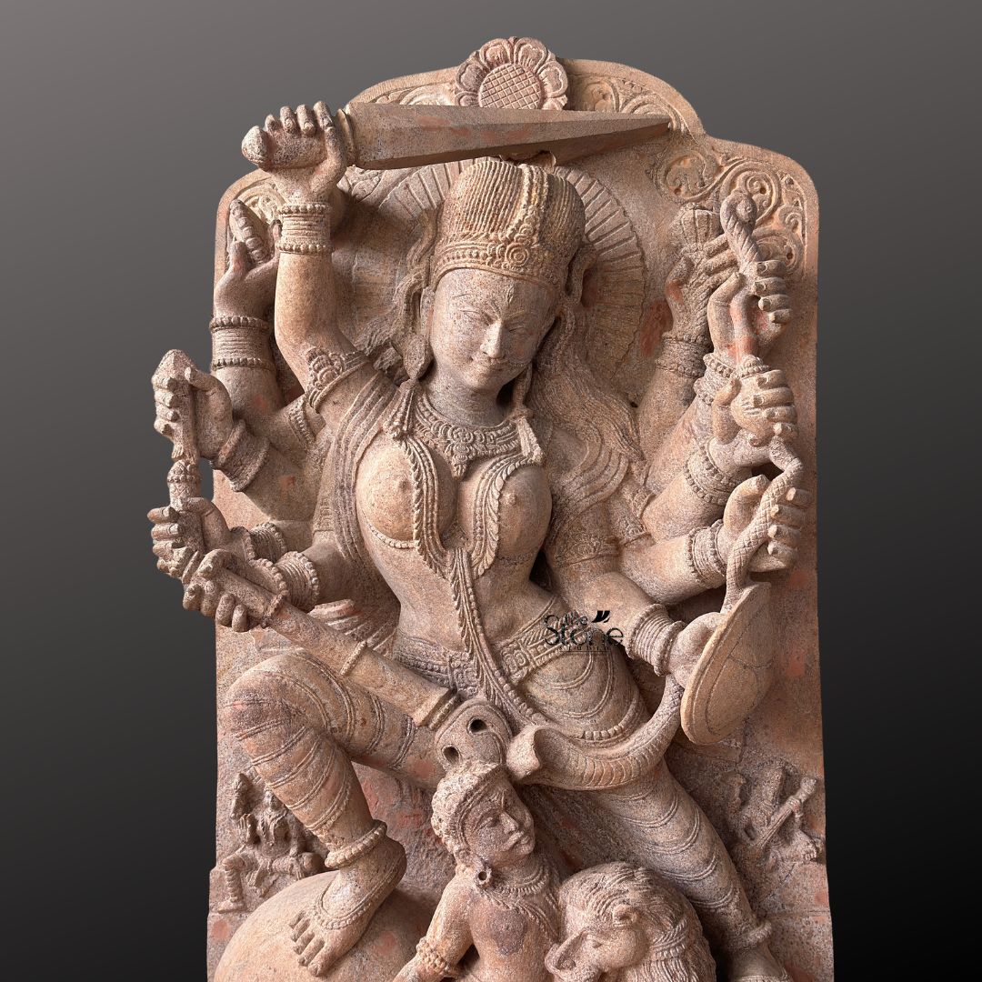 This magnificent stone sculpture carved from a warm-toned pinkish-red sandstone, depicts the Hindu goddess Durga in her powerful form as Mahishasura Mardini the slayer of the buffalo demon. The statue is a high-relief stele showcasing intricate classical Indian artistry. Durga is shown in a dynamic active posture mid-battle with her right foot planted firmly on the severed head of the buffalo demon and her left leg bent. Her primary right hand wields a sword held aloft while her primary left hand holds the tail of the demon who is emerging in human form from the buffalo's carcass. The goddess is depicted with eight arms each holding a symbolic weapon or item including a trident conch disc bow and arrow all rendered in fine detail. She wears elaborate jewelry a detailed crown and a flowing dhoti. The demon Mahishasura is shown in a contorted defeated posture beneath her foot with a lion mount attacking him from the side. The entire sculpture is framed by an ornate archway with floral and geometric carvings and the base includes small relief figures and a central plinth emphasizing the sacred nature of this Mahishasura Mardini Durga statue in Sandstone. The statue is presented against a dark neutral grey background which allows the natural texture and warm color of the sandstone to stand out. The lighting is directiona casting soft shadows that enhance the depth and three-dimensionality of the complex carving showcasing the skilled artisanship. This image is an ideal representation for an article discussing the iconography spiritual significance and cultural heritage of the Goddess Durga Stone Statue for Temple.