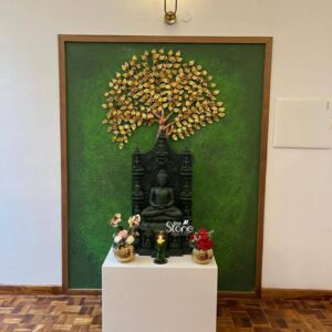 This is a beautifully composed eye-level photograph of a sacred indoor shrine featuring a striking hand-carved green stone Buddha statue as the central deity. The statue standing approximately 3 feet tall is crafted from a distinct dark green natural stone showcasing a smooth polished finish that highlights the rich varied color and fine texture of the material. The Buddha is depicted in a seated meditative Dhyana mudra postur with hands resting palms-up in the lap. He is seated upon a traditional multi-tiered green stone throne carved with intricate traditional relief patterns and figures which provides a dark dramatic contrast to the vibrant green stone of the statue. The Buddha and its throne are positioned upon a simple clean-lined polished white cube pedestal elevating the deity for reverence and display. This entire devotional setup is set against a large textured forest-green wall panel that has been intentionally painted with a rough, stucco-like finish, creating a deep, verdant backdrop that complements and harmonizes with the green stone of the statue. This green panel is framed by a clean natural wood border integrating the shrine into the room's architecture. Above the statue mounted on the green wall is a decorative metal art piece resembling a gilded Bodhi tree with a textured trunk and dense canopy of hammered gold-leaf-shaped leaves symbolizing enlightenment. The shrine is located in a home environment in Kodaikanal evidenced by the rich warm-toned wooden parquet flooring in the foreground and the clean light-colored walls of the room. On the white pedestal flanking the statue are offerings arranged to enhance the devotional atmosphere: to the left, a small polished gold vase holds delicate pink and white orchids; to the right, a matching gold vase displays vibrant red roses. In the center directly in front of the Buddha, sits a single, lit green pillar candle in a simple black holder, casting a soft, warm glow that emphasizes the three-dimensional form and natural color of the green stone carving. The overall composition presents a blend of traditional artistry and contemporary home decor.