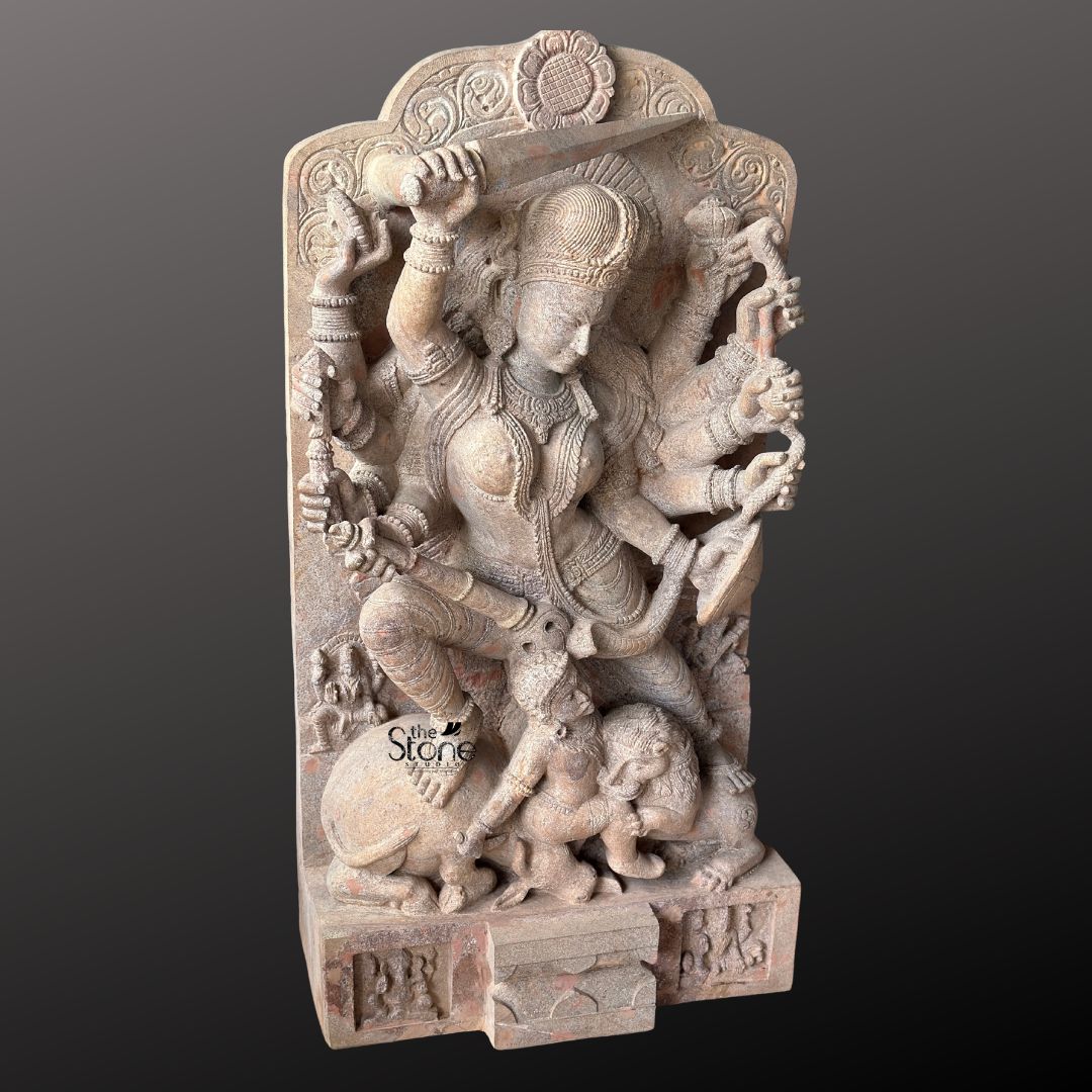 This magnificent stone sculpture expertly hand-carved from a warm, pinkish-hued sandstone, depicts the Hindu goddess Durga in her powerful form as Mahishasura Mardini the slayer of the buffalo demon. The statue is a high-relief stele showcasing the intricate skill of traditional Indian stone artistry with deep carvings that create a strong sense of form and motion. Durga is shown in a dynamic active posture mid-battle with her right foot planted firmly on the severed head of the buffalo demon and her left leg bent. Her primary right hand wields a sword held aloft while her primary left hand holds the tail of the demon who is emerging in human form from the buffalo's carcass. The goddess is depicted with numerous arms each holding a symbolic weapon or item including a trident conch disc and bow all rendered with fine detail. She wears elaborate jewelry armlets bracelets a detailed crown and a flowing dhoti all carved with delicate patterns that enhance the three-dimensional quality of the stone. The demon Mahishasura is shown in a contorted defeated posture beneath her foot with a lion mount attacking him from the side. The entire sculpture is framed by an ornate archway with floral and geometric carvings and the base includes small relief figures further emphasizing the sacred nature of this Mahishasura Mardini Durga statue in Sandstone. The statue is presented against a dark neutral grey background which allows the natural texture and warm color of the sandstone to stand out. The lighting is directional casting soft shadows that enhance the depth and three-dimensionality of the complex carving showcasing the skilled craftsmanship. This image is an ideal representation for an article discussing the iconography spiritual significance and cultural heritage of a Hand Carved Durga Maa Stone Statue.