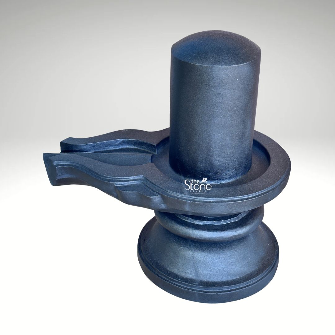 A three-quarter studio photograph captures a traditional Blackstone Shivling statue meticulously hand-carved from a single piece of dark rich blackstone. The entire murti rests on a simple, rounded pedestal base providing a stable foundation. Rising from this base is the yoni pedestal featuring a smooth perfectly centered shivling shaft. The dark polished surface of the stone absorbs and reflects light subtly emphasizing the heavy solid presence of the murti and the tactile quality of the material. The clean neutral background ensures that the viewer's attention is entirely on the sculpture's form and texture. The artistry of this hand-carved stone Shivling with yoni is evident in the fine detail and traditional iconography. The smooth cylindrical linga represents the formless aspect of Lord Shiva while the yoni base symbolizes the creative energy of Goddess Parvati. The harmonious union of these two elements is central to Hindu philosophy. To the side the intricately carved pranala extends outwards showing precise channels and traditional decorative motifs that are characteristic of fine Indian stonemasonry. The entire piece embodies a sense of solemnity and divine balance. This image serves as an exemplary representation of a Hand-Carved Stone Shivling with Yoni highlighting the specific craftsmanship and quality of the blackstone used. The photograph effectively communicates the cultural significance spiritual symbolism and physical beauty of the murti making it a valuable visual reference for devotees and collectors interested in acquiring a traditionally crafted Shivling for a home or temple shrine. The high-quality lighting enhances the deep texture of the stone showcasing the skill of the artisan in every curve and line.