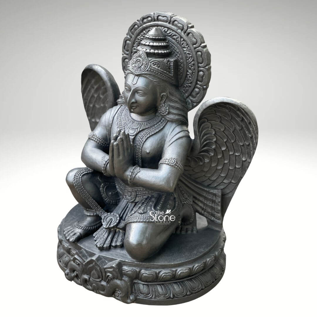 A centered three-quarter view studio photograph presents a meticulously hand-carved black stone statue of Garuda standing approximately two feet tall against a clean light grey gradient background. The statue is a unified piece of dark polished granit showcasing the stone's natural finely crystalline texture and deep uniform color. Garuda the divine eagle-man and mount of Lord Vishnu is depicted in a kneeling reverential posture balancing gracefully on his right leg while his left foot rests flat on an intricately carved circular lotus petal base. His hands are pressed together at his chest in the Anjali Mudra conveying deep devotion and respect. His serene human-like face gazes forward with a composed expression framed by elaborate traditional jewelry necklaces armlets wristlets and a tall ornate crown that is further backed by a decorative radiating prabhavali featuring a kirtimukha motif at its apex. Garuda’s iconic powerful wings are fully unfurled and spread symmetrically behind his torso, filling the upper background and exhibiting detailed feather carving that contrasts with the smooth dark surface of his body. He wears a sculpted dhoti with rich patterns and a central flowing sash all defined by precise lines and intricate detailing that highlight the sculptor's skill. The entire sculpture demonstrates exceptional craftsmanship with sharp clean edges and smooth curves that give the dark granite a fluid, lifelike quality as if it were cast in metal rather than carved from stone. The single-piece construction from durable black stone enhances its sense of ancient sanctity and spiritual presence. This image serves as an ideal visual reference for an article discussing the traditional iconography and spiritual significance of a Handcrafted Garuda Stone Statue for Temple. The detailed depiction of the dark stone, the precise execution of the prayer gesture and the impressive scale of the wings are all key elements relevant to the article's content. The two-foot height makes it a perfect size for home shrines and temple niches representing devotion protection and the divine vehicle of Vishnu. The high-quality finish and accurate proportions make this particular statue an excellent representation of a traditional Hindu devotional murti.