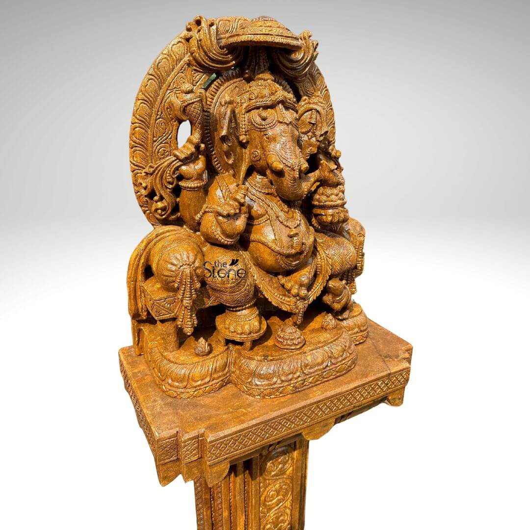 This is a focused three-quarter view photograph of a magnificent hand-carved brown sandstone Murti of Lord Ganesh elevated and displayed upon a matching ornately carved sandstone pedestal. The Murti depicts Lord Ganesh in a traditional seated posture known as Vighneshvara upon a richly carved throne. He is adorned with intricate traditional jewelry a detailed crown and garlands all carved directly from the warm textured brown sandstone. Ganesh has four arms holding symbolic attributes including an axe and a goad with his primary right hand in the Abhaya mudra of blessing. His form is robust and detailed with a serene facial expression and deep complex carvings that showcase artisanal skill. The Ganesh Murti rests upon a substantial square sandstone pedestal base which is richly carved on all visible sides with traditional Indian motifs including decorative floral patterns miniature figural reliefs and architectural elements that echo the artistry of the main sculpture. The pedestal provides a stable and elegant foundation raising the deity to a prominent height suitable for worship or prominent display as a decor piece. The entire ensemble—Murti throne and pedestal—is crafted from the same consistent brown sandstone featuring a beautiful uniform natural grain and a finely textured matte finish that emphasizes the depth and complexity of the hand-carvings. The Murti and pedestal are presented against a clean seamless light-grey studio background that fades to white isolating the object and allowing its form color and intricate details to be the sole focus. The lighting is soft even and directional illuminating the scene from the front and slightly to the side to cast gentle shadows that define the three-dimensional quality and deep reliefs of the sandstone carving. The overall composition conveys a sense of spiritual reverence traditional artistry and substantial craftsmanship inherent to this type of Indian stone sculpture.