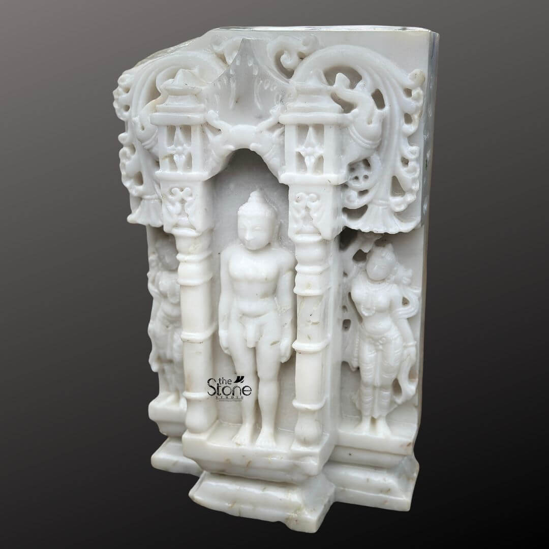 A detailed eye-level photograph captures a magnificent 18-inch Handcrafted Marble Jain Tirthankara Sculpture featuring a distinct antique-look finish. The statue depicts the Tirthankara in the meditative Kayotsarga posture standing perfectly straight and centered within a traditional ornate prabhavali also carved from the same white marble. The figure is smooth and serene with eyes half-closed in contemplation and elongated earlobes, conveying a profound sense of tranquility and detachment. The surface of the marble exhibits a subtle aged patina with natural color variations and a slightly textured feel giving it the appearance of a centuries-old devotional artifact consistent with the article's theme. The Tirthankara stands on a multi-tiered stepped marble pedestal which is embellished with carved patterns further grounding the entire divine composition. The elaborate archway frame surrounding the deity features intricate carvings of traditional motifs including stylized floral scrolls miniature shrines and two smaller flanking standing figures of yakshas or attendants on either side all rendered with fine detail in the marble. The sculpture's form is balanced and symmetrical exemplifying the high level of craftsmanship involved in creating this devotional artwork. The use of white marble while antiqued symbolizes the purity and clarity central to Jainism. This image is an exceptional representation of a Handcrafted Marble Jain Tirthankara Sculpture with a convincing antique-look finish making it an ideal centerpiece for any article discussing the beauty history and spiritual significance of such antique-style artifacts. The fine details from the delicate carvings on the archway to the serene form of the Tirthankara showcase the artisan's skill. The statue's compact 18-inch size makes it suitable for a home altar or a place of worship while its aged aesthetic adds a layer of historical reverence. The clean dark grey background ensures the white marble figure stands out with clarity allowing every nuance of the carving to be appreciated.