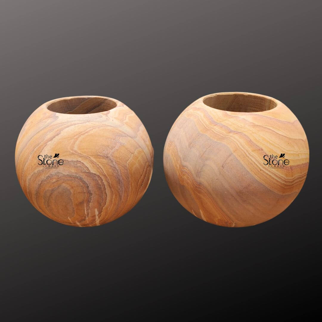 A studio photograph presents a pair of identical spherical sandstone planter pots arranged side-by-side against a softly graduated dark grey background. Each pot is hand-carved from a single block of natural sandstone showcasing a rich warm honey-gold base color interwoven with distinctive flowing layered strata patterns in shades of cream beige and light brown. This unique natural veining gives each piece a one-of-a-kind artisanal appearance. The pots are perfectly spherical with a smooth tactile matte finish that emphasizes the inherent beauty of the stone. The top of each sphere is hollowed out to form a perfectly circular deep basin approximately half the diameter of the pot designed to hold a plant and soil. The rim of the opening is clean and smooth leading down into the planting chamber. The texture of the sandstone is clearly visible exhibiting a fine-grained slightly porous quality that is both durable and aesthetically pleasing. The substantial weight and solid form of the pots convey a sense of stability and quality craftsmanship. These handcrafted sandstone garden planters are specifically designed to be a sophisticated and organic addition to any home decor or garden setting. The earthy colors and flowing patterns of the stone complement a wide range of plant life from succulents and cacti to small flowering plants or ferns. The spherical shape is a timeless and elegant design choice that brings a soft natural element to modern or traditional spaces. The durability of the sandstone makes them suitable for both indoor and outdoor use ensuring they remain a beautiful and functional piece for years to come. Their artisanal quality and natural material highlight a commitment to sustainable and elegant living.