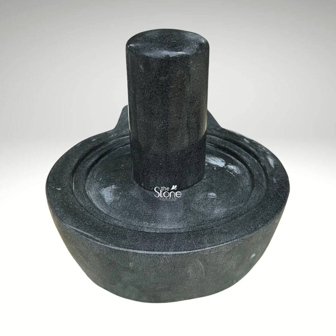 A centered eye-level studio photograph presents a meticulously hand-carved black stone Shiva Lingam with its associated Yoni base set against a clean light grey gradient background. The complete murti is crafted from a single block of dense dark black granite showcasing the natural finely crystalline texture and deep uniform color of the stone. The central element is the cylindrical Shiva Lingam representing the formless aspect of Lord Shiva which rises smoothly and vertically from the center of the circular Yoni pedestal. The Lingam's surface is highly polished creating a subtle sheen that reflects ambient light and contrasts with the matte finish of the surrounding elements. The Yoni base symbolizing the feminine creative energy of Goddess Parvati is a circular basin-like structure surrounding the base of the Lingam. It features a clearly defined raised circular rim and a well-proportioned projecting spout to the left side designed to channel ceremonial offerings of water or milk away from the deity. The entire Yoni base is meticulously carved from the same dark black stone with precise concentric rings enhancing its form and balance. The carving exhibits traditional craftsmanship with smooth flowing curves and precise edges that give the Shivling and Yoni a harmonious and integrated appearance conveying a sense of ancient sanctity. This image serves as a prime visual reference for an article discussing the traditional iconography and spiritual significance of a Handcrafted Shiva Lingam in Black Stone. The detailed depiction of the dark granite the specific shape of the Yoni and the presence of the spout are all key elements relevant to the article's content. The single-piece construction makes it a sought-after item for home shrines and temples representing the union of masculine and feminine energies in Hindu philosophy. The high-quality finish and accurate proportions make this particular statue an excellent representation of a traditional devotional murti.