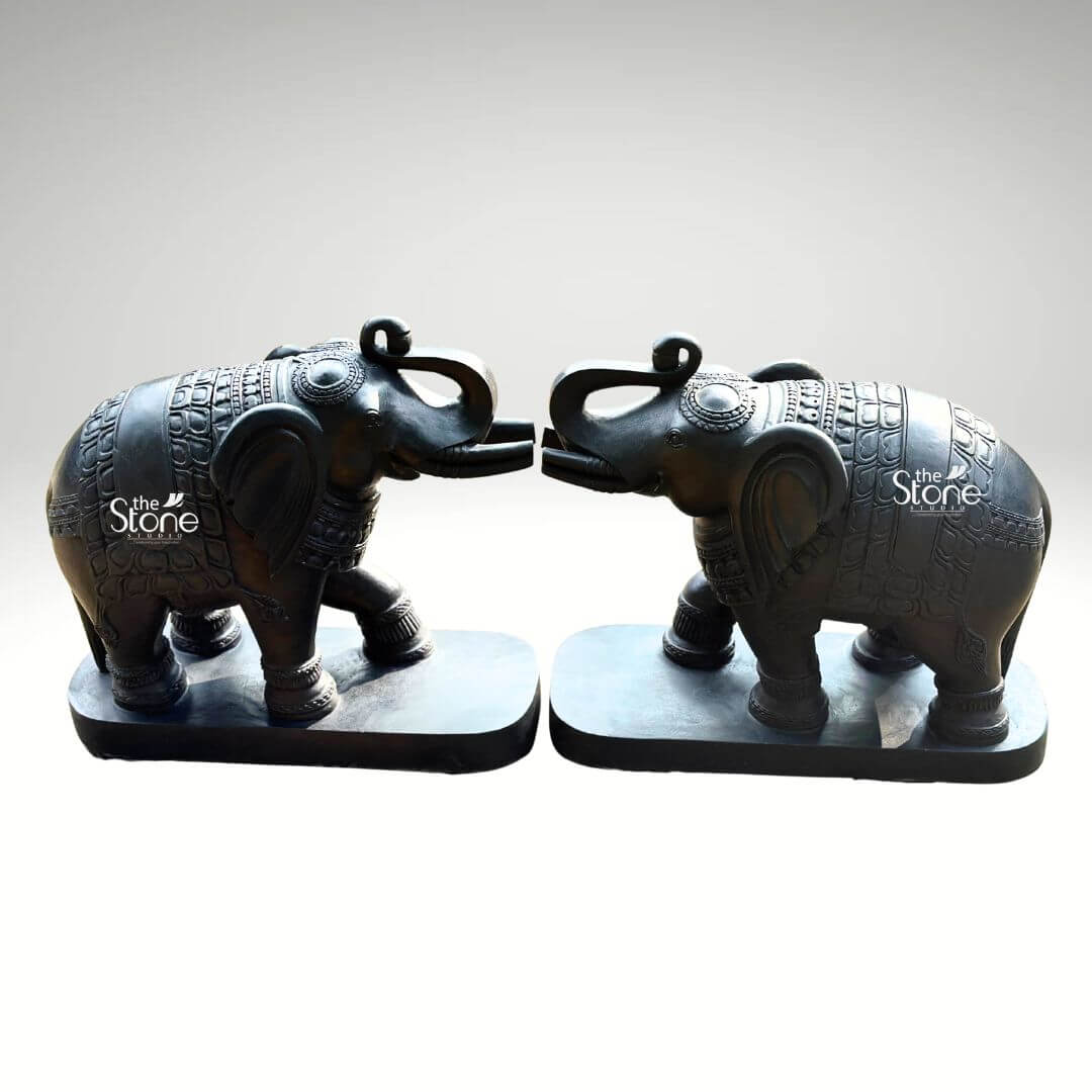 A studio product photograph captures a magnificent symmetrical pair of 26-inch blackstone elephant statues hand-carved from a single piece of dark rich stone. The statues are positioned facing each other with their raised trunks touching gently in an affectionate gesture often associated with greeting good fortune and strength. Each elephant is mounted on a simple unadorned black rectangular base which grounds the substantial figures. The entire composition is presented against a clean neutral light-grey background allowing the dark stone to stand out with a commanding presence. The detailed artistry of the statues showcases exceptional traditional Indian craftsmanship. The smooth polished surface of the blackstone reveals subtle natural variations in the stone's texture while reflecting ambient studio light to accentuate the deep charcoal color. The elephants are richly adorned with finely etched patterns that cover their bodies forming intricate designs for ornamental blankets draped over their backs as well as decorative details on their ears headpieces and legs. The precision of the hand-carving is evident in the textured patterns of the blankets and the defined wrinkles on the elephants' trunks and legs creating a highly detailed and tactile appearance. This 26-inch blackstone Elephant Pair statue set is a perfect illustration of a Handcrafted Stone Elephant Pair Statue embodying the cultural significance and spiritual symbolism of such pieces. The powerful elegant stone material and the specific pose of the elephants with interlocked trunks make it an ideal decorative element for a grand entryway, living room or office believed to bring positive energy and prosperity. The high-quality photograph clearly conveys the scale texture and intricate detail of the pair highlighting the unique artisanal value that makes such murtis a treasured addition to any home decor.
