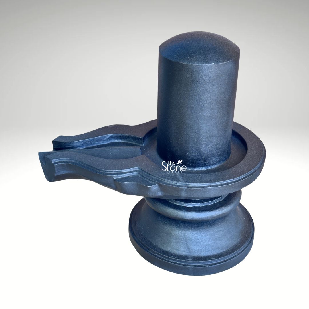 A three-quarter studio photograph captures a traditional Blackstone Shivling statue meticulously hand-carved from a single piece of dark rich blackstone. The entire murti rests on a simple, rounded pedestal base which provides a stable foundation. Rising from this base is the yoni pedestal featuring a perfectly centered smooth shivling shaft. The dark polished surface of the stone absorbs and reflects light subtly emphasizing the heavy solid presence of the murti and the tactile quality of the material. The clean neutral background ensures that the viewer's attention is entirely on the sculpture's form texture and traditional iconography. The artistry of this handmade Shivling is evident in the fine detail and traditional iconography. The smooth cylindrical linga represents the formless aspect of Lord Shiva while the yoni base symbolizes the creative energy of Goddess Parvati. The harmonious union of these two elements is central to Hindu philosophy. A key feature and the focus of the article is the intricately carved jaladhari that extends outwards from the yoni pedestal. It shows precise channels and traditional decorative motifs a testament to the artisan's skill in fine Indian stonemasonry designed to catch and channel ritual offerings of water milk and other liquids. This black stone Shivling with jaladhari is an exemplary representation of a handmade devotional object suitable for a home shrine or temple. The precise craftsmanship ensures that the murti is not only a spiritual focal point but also a work of art that embodies a sense of solemnity and divine balance. The high-quality photograph effectively communicates the rich texture of the stone and the artisanal value of the piece making it a valuable visual reference for individuals interested in acquiring a traditionally crafted Mahadev Shivling for their personal worship.