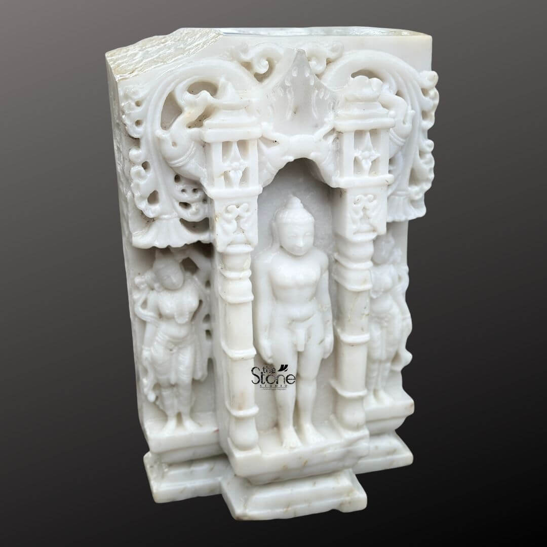 A detailed eye-level photograph captures a magnificent 18-inch Jain Bhagwan Marble Statue meticulously hand-carved and featuring a distinct antique-look finish designed for a temple. The statue depicts the Tirthankara in the meditative Kayotsarga posture standing perfectly straight and centered within a traditional ornate prabhavali also carved from the same white marble. The figure is smooth and serene with eyes half-closed in contemplation and elongated earlobes conveying a profound sense of tranquility and detachment. The surface of the marble exhibits a subtle aged patina with natural color variations and a slightly textured feel giving it the appearance of a centuries-old devotional artifact consistent with the article's theme of an antique-look statue. The Tirthankara stands on a multi-tiered stepped marble pedestal which is embellished with carved patterns further grounding the entire divine composition. The elaborate archway frame surrounding the deity features intricate carvings of traditional motifs including stylized floral scrolls miniature shrines and two smaller flanking standing figures of yakshas or attendants on either side all rendered with fine detail in the marble. The sculpture's form is balanced and symmetrical exemplifying the high level of craftsmanship involved in creating this devotional artwork. The use of white marble while antiqued symbolizes the purity and clarity central to Jainism. This image is an exceptional representation of a Jain Bhagwan Marble Statue with a convincing antique-look finish making it an ideal centerpiece for any article discussing the beauty history and spiritual significance of such antique-style artifacts destined for a temple. The fine details from the delicate carvings on the archway to the serene form of the Tirthankara, showcase the artisan's skill. The statue's compact 18-inch size makes it suitable for a home altar or a place of worship while its aged aesthetic adds a layer of historical reverence. The clean dark grey background ensures the white marble figure stands out with clarity, allowing every nuance of the carving to be appreciated.
