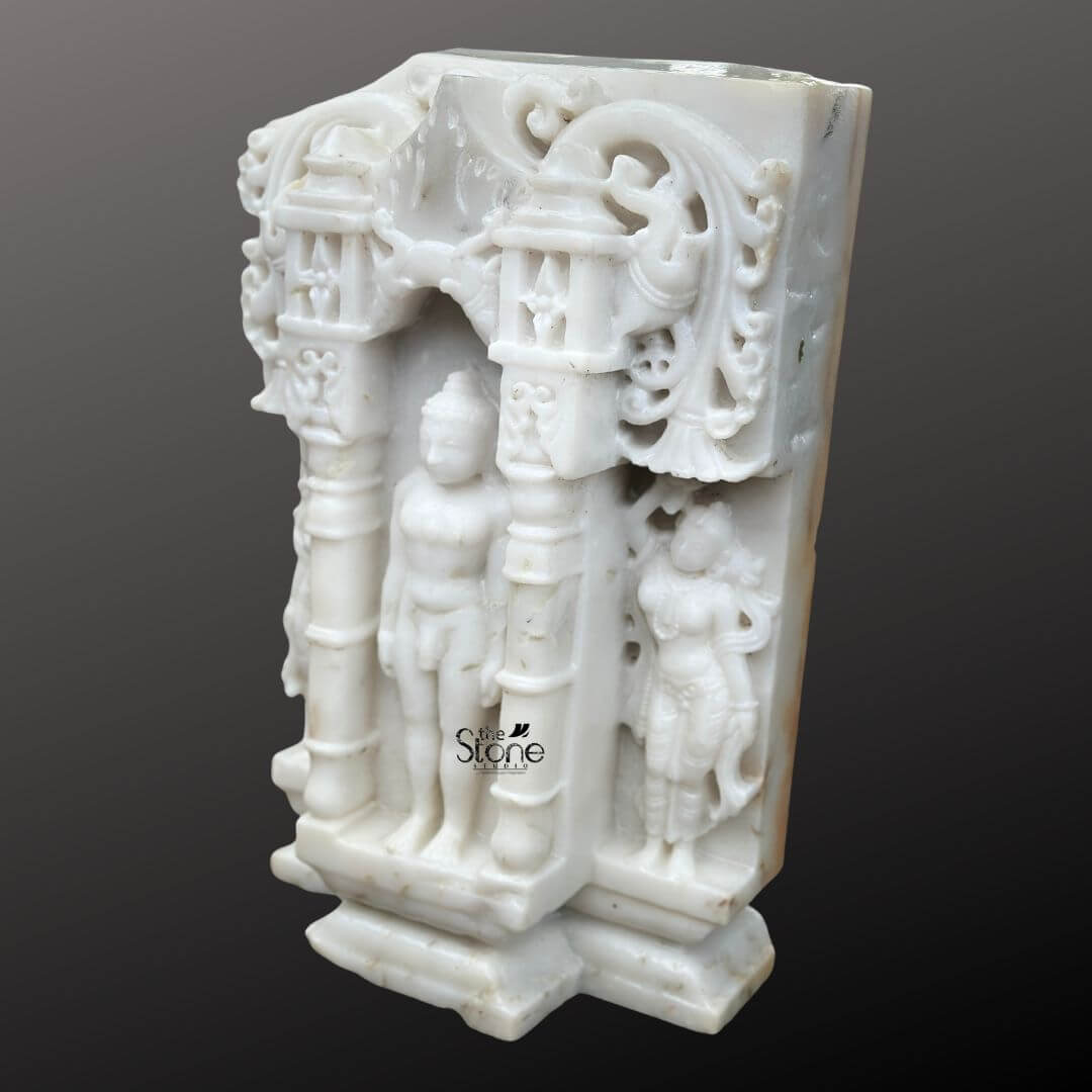 A detailed eye-level photograph captures a magnificent 18-inch Jain Tirthankar Idol meticulously hand-carved from pristine white marble and featuring a distinct antique-look finish. The central Tirthankar figure is depicted in the meditative Kayotsarga posture standing perfectly straight with arms held closely but not touching the body embodying absolute detachment and tranquility. The figure is smooth and serene with eyes half-closed in contemplation and elongated earlobes. The statue is centered within a traditional ornate prabhavali which is also carved from the same white marble and designed to resemble a miniature temple consistent with the article's theme. The Tirthankar stands on a multi-tiered stepped marble pedestal which is embellished with carved patterns. The elaborate temple-design archway surrounding the deity features intricate carvings of traditional motifs including stylized floral scrolls miniature shrines and two smaller flanking standing figures of yakshas or attendants on either side. The entire composition is balanced and symmetrical exemplifying the high level of craftsmanship involved in creating this devotional artwork. The surface of the white marble exhibits a subtle aged patina with natural color variations and a slightly textured feel giving it the appearance of a centuries-old devotional artifact. This image is an exceptional representation of a Jain Tirthankar Idol with Temple Design showcasing the rich artistic and spiritual heritage of Jain iconography. The fine details from the delicate carvings on the miniature temple archway to the serene form of the Tirthankar demonstrate the artisan's skill. The use of white marble symbolizes purity and clarity, core tenets of Jainism making the statue a fitting centerpiece for any article discussing the significance and beauty of such artifacts. The clean dark grey background ensures the white marble figure stands out with clarity allowing every nuance of the carving to be appreciated.