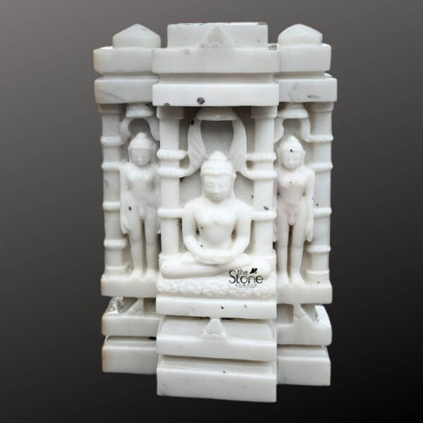 A centered eye-level studio photograph presents a meticulously hand-carved white marble statue depicting a Jain Tirthankara triad within a miniature temple archway standing approximately 17 inches tall against a dark neutral gradient background. The central figure is a seated Tirthankara presumably Lord Mahavira in a meditative lotus posture on a decorated lotus pedestal. His hands are in the dhyana mudra in his lap. He is depicted with a calm serene expression gently closed eyes and tightly curled hair in the traditional ushnisha. The central Tirthankara is enshrined within an elaborate multi-tiered marble temple arch or prabhavali. This arch is composed of intricate architectural elements including miniature pillars carved panels and a decorative upper frieze culminating in a stylized kirtimukha motif at the apex. Flanking the central seated figure standing in a rigid upright posture are two smaller Tirthankara figures on either side also carved from the same white marble and integrated into the architectural framework of the shrine. The entire piece is a unified carving of high-quality white marble showcasing the stone's natural subtle translucency and smooth polished finish. The architectural details of the temple arch are sharp and precise with delicate floral and geometric patterns defining the pillars arches and decorative elements. The three Tirthankara figures are smooth and sculpted with idealized proportions conveying a sense of spiritual tranquility and timeless devotion. The lighting is soft and even highlighting the intricate carvings and the cool luminous quality of the marble. This image serves as a prime example of traditional Jain iconography showcasing a revered Tirthankara triad in an elaborate marble temple arch suitable for home or temple shrines. The 17-inch size and detailed carving make it a significant piece of devotional art.