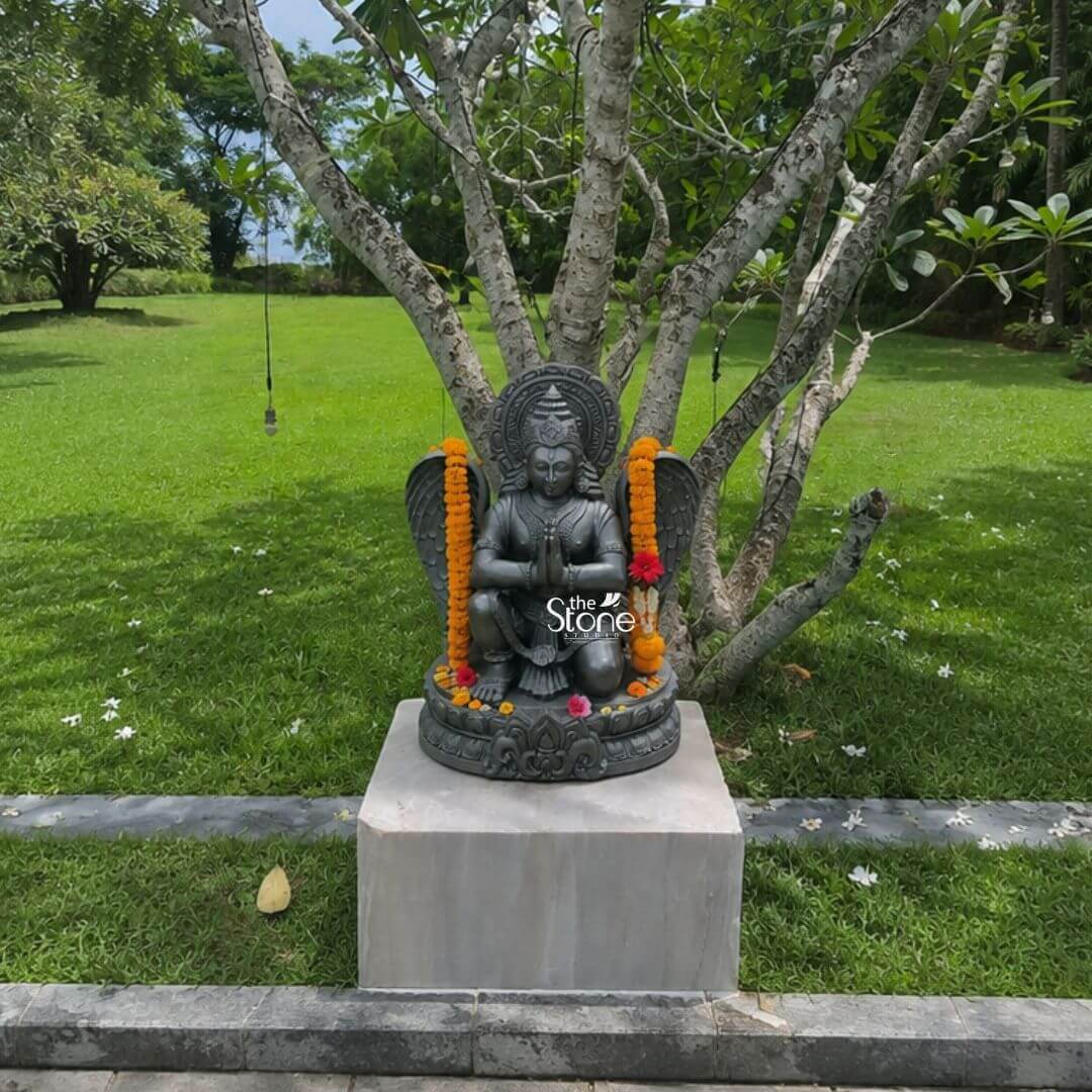 A centered eye-level photograph captures a meticulously hand-carved black stone statue of Garuda, standing approximately two feet tall installed outdoors in a serene well-manicured garden. The statue is elevated on a simple solid light-grey stone plinth which is positioned at the edge of a grey stone pathway that borders a lush green lawn. The background features a large mature tree with gnarled, light-colored branches and abundant green foliage creating a natural canopy. The entire scene is bathed in bright natural daylight casting soft defined shadows that highlight the intricate details and dark polished texture of the granite sculpture. The Garuda statue is depicted in a kneeling reverential posture with its right knee on the ground and its left leg bent balancing gracefully on an intricately carved circular lotus petal base. Its hands are pressed together at its chest in the Anjali Mudra conveying deep devotion and respect. Garuda’s human-like face gazes forward with a composed expression framed by elaborate traditional jewelry necklaces armlets wristlets and a tall ornate crown that is further backed by a decorative radiating prabhavali. The deity is adorned with fresh garlands of vibrant orange marigolds draped over its shoulders and a few loose pink and red flowers scattered at its feet on the lotus base. Garuda’s iconic powerful wings are fully unfurled and spread symmetrically behind its torso exhibiting detailed feather carving that contrasts with the smooth, dark surface of its body. It wears a sculpted dhoti with rich patterns and a central flowing sash all defined by precise lines and intricate detailing that highlight the sculptor's skill. The sculpture demonstrates exceptional craftsmanship with sharp clean edges and smooth curves that give the dark granite a fluid lifelike quality as if it were cast in metal rather than carved from stone. The single-piece construction from durable black stone enhances its sense of ancient sanctity and spiritual presence within the tranquil garden environment. This image serves as an ideal visual reference for an article discussing the traditional iconography and spiritual significance of a Kneeling Garuda Statue with Folded Hands. The detailed depiction of the dark stone the precise execution of the prayer gesture the elaborate jewelry and the impressive scale of the wings are all key elements relevant to the article's content. The two-foot height makes it a perfect size for home shrines and temple niches representing devotion protection and the divine vehicle of Vishnu. The natural outdoor garden setting demonstrates a common and culturally appropriate way to enshrine such a deity making the image highly relevant for those seeking inspiration for their own sacred space. The detailed carving and balanced proportions of this particular Garuda make it an excellent representation of a traditional Hindu devotional murti.