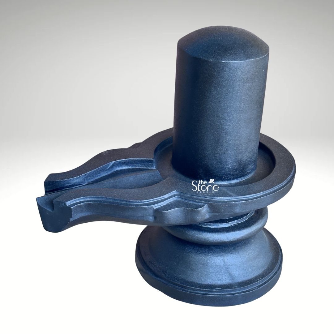 A three-quarter studio photograph captures a substantial Blackstone Shivling statue meticulously carved and polished to a deep charcoal-black finish. The murti rests on a simple rounded pedestal base which provides a stable foundation. Rising from the base is the cylindrical yoni pedestal featuring a perfectly centered smooth shivling shaft. To the side the intricately carved pranala or jaladhari extends outwards showing precise channels and traditional decorative motifs that are characteristic of fine Indian stonemasonry. The entire form has a smooth tactile quality with the dark stone absorbing and reflecting light in a way that emphasizes its heavy solid presence. This large black stone Shivling is an exceptional example of traditional temple sculpture designed to be a commanding focal point in a place of worship. The size and scale of the piece convey a sense of grandeur and spiritual power, making it appropriate for the article's focus on temple installations. The precise carving of the yoni and the smooth cylindrical linga represent the harmonious union of Shiva and Shakti a central concept in Hindu philosophy. The dark rich color of the stone adds to the solemn and divine atmosphere that such a murti is intended to evoke within a temple's garbhagriha. The image showcases the specific details and craftsmanship of this large Blackstone Shivling statue highlighting its suitability for significant temple projects. The clean neutral background ensures that the viewer's attention is entirely on the sculpture's form texture, and traditional iconography. The photograph effectively communicates the quality of the stone and the artisan's skill providing a valuable visual reference for architects temple committees and devotees interested in acquiring a monumental Shivling for a temple setting.