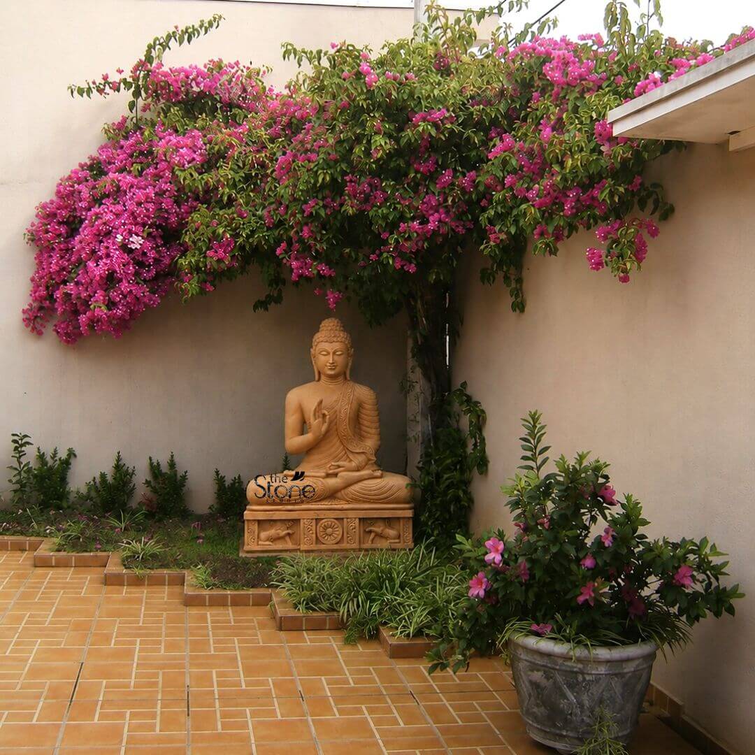 A tranquil eye-level photograph captures a magnificent 4-foot tall Buddha statue as the focal point of a peaceful outdoor garden courtyard. The statue is expertly hand-carved from warm, textured brown sandstone showcasing a rich natural finish that harmonizes with the environment. It is seated in the traditional vajra lotus position on a carved pedestal embodying deep meditation and peace. The figure is draped in intricately carved monastic robes with the fabric folds and a decorative patterned border clearly defined on the left shoulder. The Buddha's face is serene with eyes half-closed in contemplation and a gentle compassionate smile seated against a textured light-beige wall. The statue is perfectly situated under the protective canopy of a large vibrant bougainvillea plant that cascades down from above creating a natural arch of brilliant fuchsia and pink flowers that frame the figure. This botanical setting enhances the spiritual ambiance making the statue appear as if it is meditating in a sacred grove. The ground in the foreground consists of warm terracotta-colored brick pavers arranged in a geometric pattern leading the eye toward the statue. Surrounding the base of the statue's pedestal are various smaller green plants and groundcovers and to the right a large weathered stone planter holds more blooming pink flowers adding depth and texture to the garden design. This image perfectly illustrates how a large 4-foot outdoor Buddha statue in brown sandstone can transform a simple garden corner into a sanctuary of calm. The substantial height of the statue makes it an imposing and inspiring presence while the natural brown sandstone weathers gracefully integrating with the organic elements like the bougainvillea and surrounding plants. The composition is balanced and inviting suggesting a perfect spot for quiet reflection and meditation. The directional daylight highlights the exquisite details of the carving from the curls of the ushnisha to the figures on the base against the soft colors of the flowers and walls.