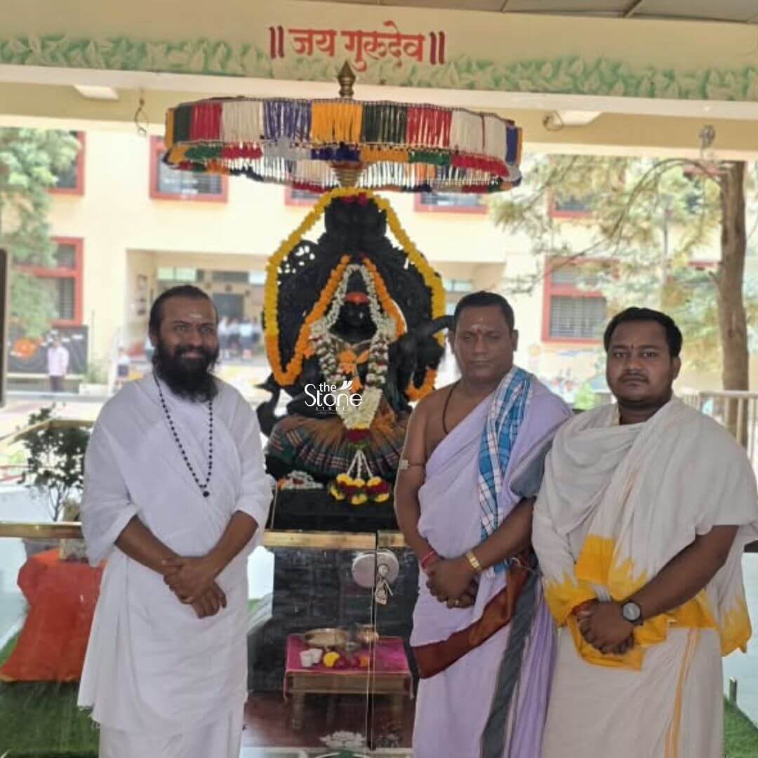 A centered full-view photograph captures a significant moment on a school campus where a large Saraswati Murti in Black Stone serves as the spiritual and cultural centerpiece. The 7-foot tall statue hand-carved from a single imposing block of deep dark blackstone is installed on a multi-tiered stone pedestal base that elevates it prominently. Goddess Saraswati the Hindu deity of knowledge music arts wisdom and learning is depicted in a classic seated posture with her form exhibiting exquisite detail. Fine carvings delineate her kiritamukuta multiple necklaces armlets wristlets and the flowing delicately rendered patterns of her dhoti all chiseled with precision into the dark stone contrasting beautifully with the bright outdoor light. The statue is richly adorned for worship draped in multiple traditional garlands. A prominent long garland of fragrant white jasmine and rose petals hangs around her neck layering over a smaller vibrant garland of yellow marigolds and orange marigolds that frames her face and torso. Her serene expression conveys deep wisdom and peace, with downcast eyes. Her four arms hold traditional attributes: her upper right hand holds a japamala and her upper left hand holds a book symbolizing knowledge. Her two main lower hands are depicted in the act of playing the veena a string instrument which she holds across her lap. A traditional colorful umbrella is mounted above her head and a small puja altar with ritual items is positioned in front of the pedestal. The statue's installation is surrounded by three individuals who provide a sense of scale and context. To the left a man in white robes with a long beard stands with hands clasped and to the right two other men in traditional Hindu attire stand respectfully. The background features a school building with large windows and some surrounding trees firmly placing the murti within an educational environment. The statue's monumental size ensures it serves as a commanding and inspiring focal point embodying the cultural and spiritual values of learning and artistic pursuit making it an appropriate and respectful piece for a school campus. The detailed carving and the durable blackstone material ensure it will be a cherished and long-lasting symbol of knowledge for generations of learners.