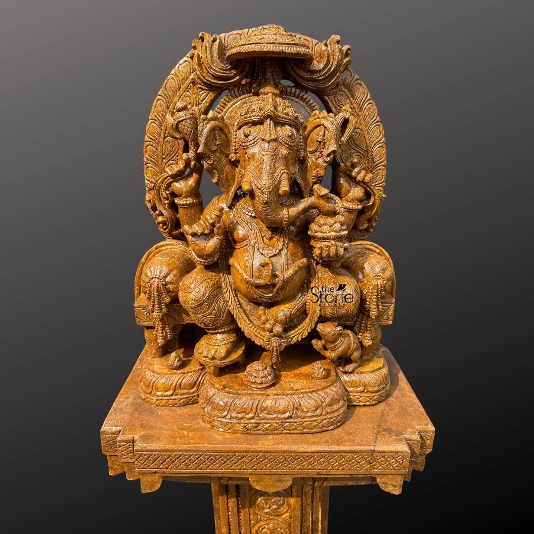 This is a full-view photograph of a magnificent hand-carved brown sandstone statue of Lord Ganesha standing approximately five feet tall and designed as a grand centerpiece for a home entrance or foyer. The statue is crafted from a warm rich-toned brown sandstone featuring a finely textured matte finish that emphasizes the intricate artisanal craftsmanship. Ganesha is depicted in a traditional seated posture known as Vighneshvara on a richly carved multi-tiered throne. He is adorned with elaborate traditional jewelry a detailed crown and garlands all carved directly from the stone with deep reliefs. Ganesha has four arms holding symbolic attributes including an axe and a goad with his primary right hand in the Abhaya mudra of blessing. The Ganesha statue rests upon a substantial square sandstone pedestal base which is richly carved on all visible sides with traditional motifs including decorative floral patterns and miniature figural reliefs that echo the art style of the main sculpture. The pedestal provides a stable and elegant foundation elevating the deity to a prominent height suitable for display in an entranceway. The entire ensemble—statue throne and pedestal—is crafted from the same consistent brown sandstone featuring a beautiful uniform natural grain that adds to the sculptural quality and visual appeal. The statue and pedestal are presented against a seamless, dark grey studio background that fades to black isolating the object and allowing its form color and intricate details to be the sole focus. The lighting is soft even and directional illuminating the scene from the front and slightly to the side to cast gentle shadows that define the three-dimensional quality and deep reliefs of the sandstone carving. The overall composition conveys a sense of spiritual reverence traditional artistry and substantial craftsmanship inherent to this type of large Indian stone sculpture making it a commanding and welcoming presence for any entrance.