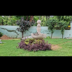 This is a centered eye-level photograph capturing a substantial 54-inch tall sandstone water fountain positioned as the central focal point within a landscaped residential garden. The fountain is hand-carved from a warm light-beige natural sandstone featuring a classic tiered design. It rests in a circular well-maintained patch of green lawn immediately surrounded by a dense thriving bed of dark purple and green foliage plants that provide a striking color contrast to the stone. The fountain itself is composed of a large shallow basin supported by a sturdy decorative pedestal base. Rising prominently from the center of the main basin is a multi-tiered central column adorned with intricate traditional carved mouldings and a fluted midsection. This central spire culminates in a smooth perfectly spherical finial at the very top from which water is actively flowing creating a gentle shimmering cascade that trickles down the carved tiers and into the basin below. The stone's finely textured matte surface highlights the artisanal craftsmanship and the rich natural grain of the sandstone making it a durable and elegant piece of garden decor. The fountain is set against a light-grey garden wall which features a subtle decorative trellis pattern and is bordered by various mature green trees and bushes creating a lush secluded background. To the left a pile of soil and a white garden chair suggest ongoing garden projects or maintenance near the fence line. To the right a hose is visible on the ground indicating practical garden upkeep. Natural daylight filters through the canopy of the surrounding trees casting soft shadows and illuminating the varied textures of the stone foliage, and water. The overall composition presents the fountain as a harmonious and tranquil addition to the outdoor space.