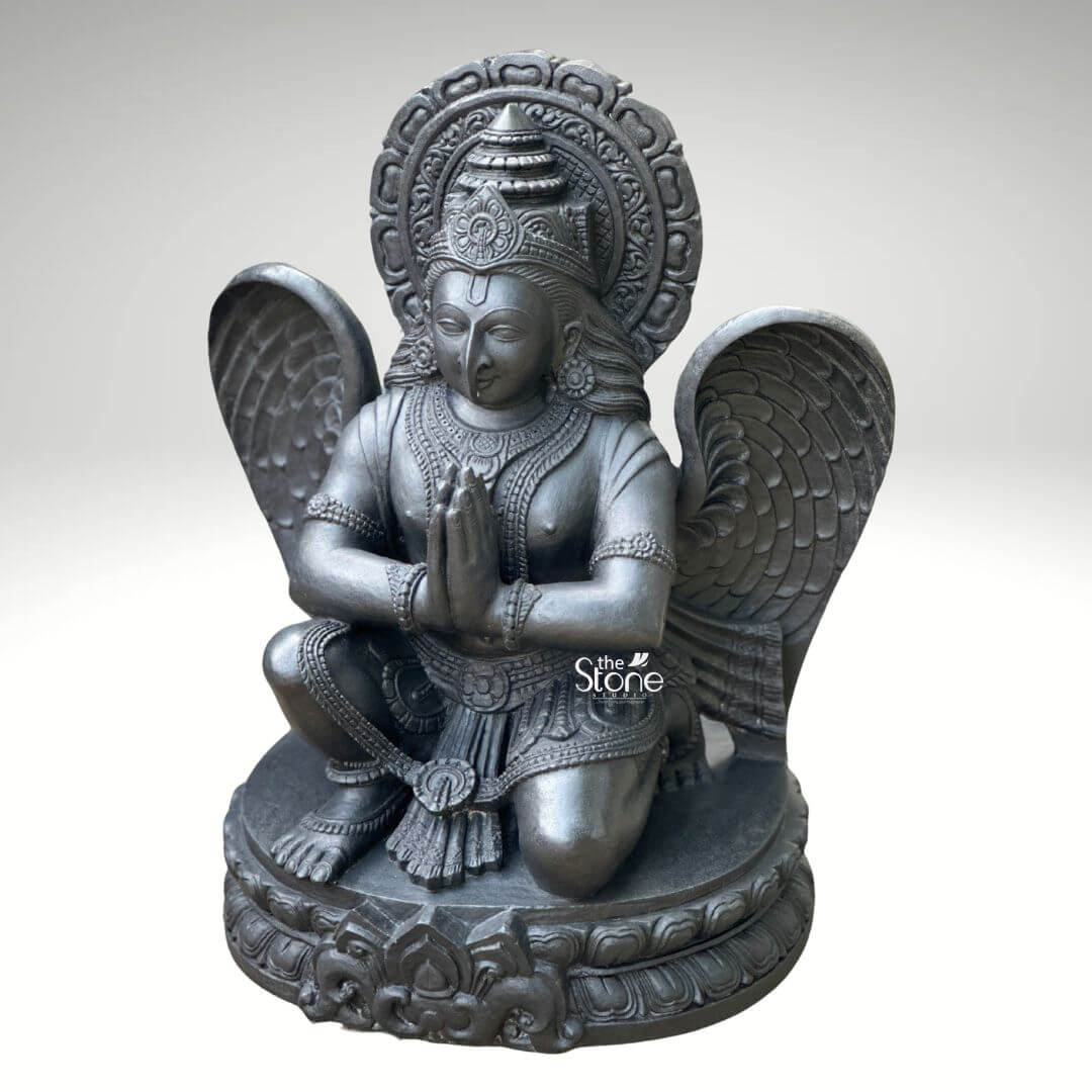 A centered eye-level studio photograph presents a meticulously hand-carved black stone statue of Lord Garuda, standing approximately two feet tall against a clean light grey gradient background. The statue is a unified piece of dark polished granite showcasing the stone's natural finely crystalline texture. Garuda the divine eagle-man and mount of Lord Vishn is depicted in a kneeling, reverential posture balancing gracefully on his right leg while his left foot rests flat on an intricately carved, circular lotus petal base. His hands are pressed together at his chest in the Anjali Mudra conveying deep devotion and respect. His serene human-like face gazes forward with a composed expression framed by elaborate traditional jewelry necklaces armlets wristlets and a tall ornate crown that is further backed by a decorative radiating prabhavali featuring a kirtimukha motif at its apex. Garuda’s iconic, powerful wings are fully unfurled and spread symmetrically behind his torso, filling the upper background and exhibiting detailed feather carving that contrasts with the smooth dark surface of his body. He wears a sculpted dhoti with rich patterns and a central flowing sash all defined by precise lines and intricate detailing that highlight the sculptor's skill. The entire sculpture demonstrates exceptional craftsmanship with sharp clean edges and smooth curves that give the dark granite a fluid, lifelike quality as if it were cast in metal rather than carved from stone. The single-piece construction from durable black stone enhances its sense of ancient sanctity and spiritual presence. This image serves as an ideal visual reference for an article discussing the traditional iconography and spiritual significance of a Lord Garuda Murti in Black Stone. The detailed depiction of the dark stone the precise execution of the prayer gesture and the impressive scale of the wings are all key elements relevant to the article's content. The two-foot height makes it a perfect size for home shrines and temple niches representing devotion protection and the divine vehicle of Vishnu. The high-quality finish and accurate proportions make this particular statue an excellent representation of a traditional Hindu devotional murti.