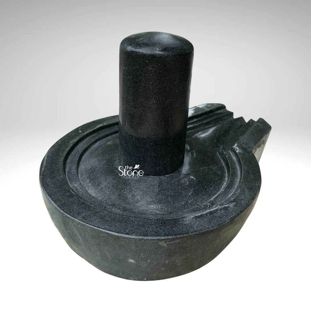 A centered eye-level studio photograph presents a meticulously hand-carved Lord Shiva Shivling statue complete with its Yoni base set against a clean light grey gradient background. The entire sacred artifact is carved from a single block of dense dark black granite showcasing the stone's natural finely crystalline texture and deep rich color. The central element is the cylindrical Shivling representing the formless aspect of Lord Shiva, which rises smoothly and vertically from the center of the circular Yoni pedestal. The top of the Shivling is gently domed and its surface is highly polished reflecting ambient light and creating a subtle sheen that contrasts with the matte finish of the surrounding elements. The Yoni base symbolizing the feminine creative energy of Goddess Parvati, is a circular basin-like structure surrounding the base of the Shivling. It features a clearly define raised circular rim and a well-proportioned projecting spout to the right side designed to channel ceremonial offerings of water or milk away from the deity. The entire Yoni base is also meticulously carved from the same dark black stone with precise concentric rings enhancing its form and balance. The carving exhibits traditional craftsmanship with smooth flowing curves and precise edges that give the Shivling and Yoni a harmonious and integrated appearance conveying a sense of ancient sanctity. This image serves as a prime visual reference for an article discussing the traditional iconography spiritual significance and proper selection of a Lord Shiva Shivling for home shrines and temples. The detailed depiction of the dark granite the specific shape of the Yoni and the presence of the spout are all key elements relevant to the article's content. The single-piece construction from durable black granite makes it a sought-after item for daily worship and ritualistic abhishekam. The high-quality finish and accurate proportions make this particular statue an excellent representation of a traditional Hindu devotional murti suitable for creating a sacred space.