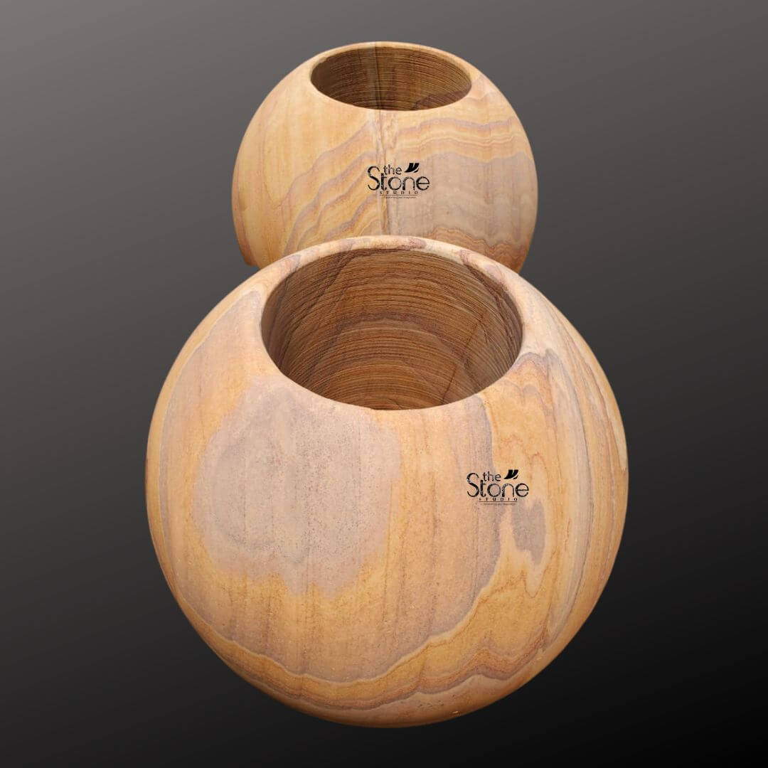 A high-angle studio photograph presents a pair of luxurious spherical sandstone planter pots stacked vertically to create an eye-catching sculptural form. The pots are crafted from a single piece of natural fine-grained sandstone exhibiting a rich warm honey-gold base color interwoven with distinctive flowing layered strata patterns in shades of cream beige and light brown. This unique natural veining resembling a topographic map or wood grain gives each pot a one-of-a-kind artisanal appearance. The surface of the stone is hand-polished to a smooth tactile matte finish that emphasizes its inherent organic beauty. The top pot is a perfect sphere slightly smaller in diameter than the lower one and is hollowed out to form a circular basin. The lower pot larger and more substantial sits directly beneath it also hollowed to create a larger planting chamber. The thickness of the sandstone wall is evident at the rim of each opening conveying a sense of durability and quality craftsmanship. The overall stacked composition is stable and elegant designed to be a prominent focal point. The substantial weight and solid form of the pots convey a sense of luxury and artisanal quality. These luxury natural stone planter pots are specifically designed to be a sophisticated and organic addition to any home decor or garden setting. The earthy colors and flowing patterns of the stone complement a wide range of plant life from succulents and cacti to small flowering plants or ferns. The spherical shape is a timeless and elegant design choice that brings a soft natural element to modern or traditional spaces. The durability of the sandstone makes them suitable for both indoor and outdoor use ensuring they remain a beautiful and functional piece for years to come. Their artisanal quality unique natural material and elegant form highlight a commitment to sustainable luxury and refined living.